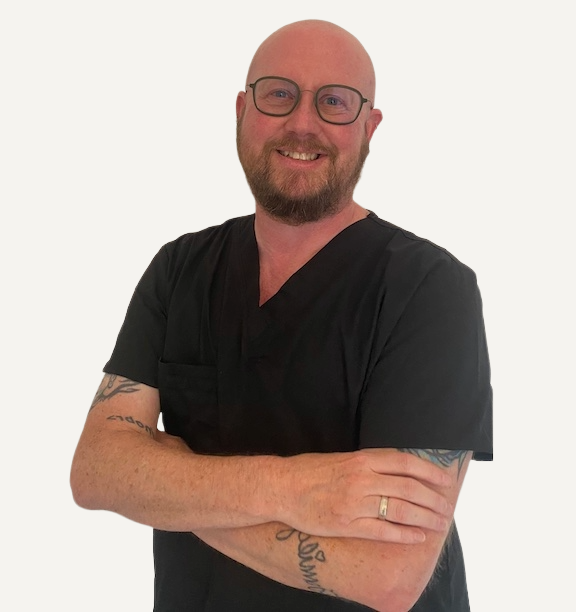 Bear smiling with glasses, beard, tattoos, wearing black medical scrubs, standing with arms crossed against a white background.
