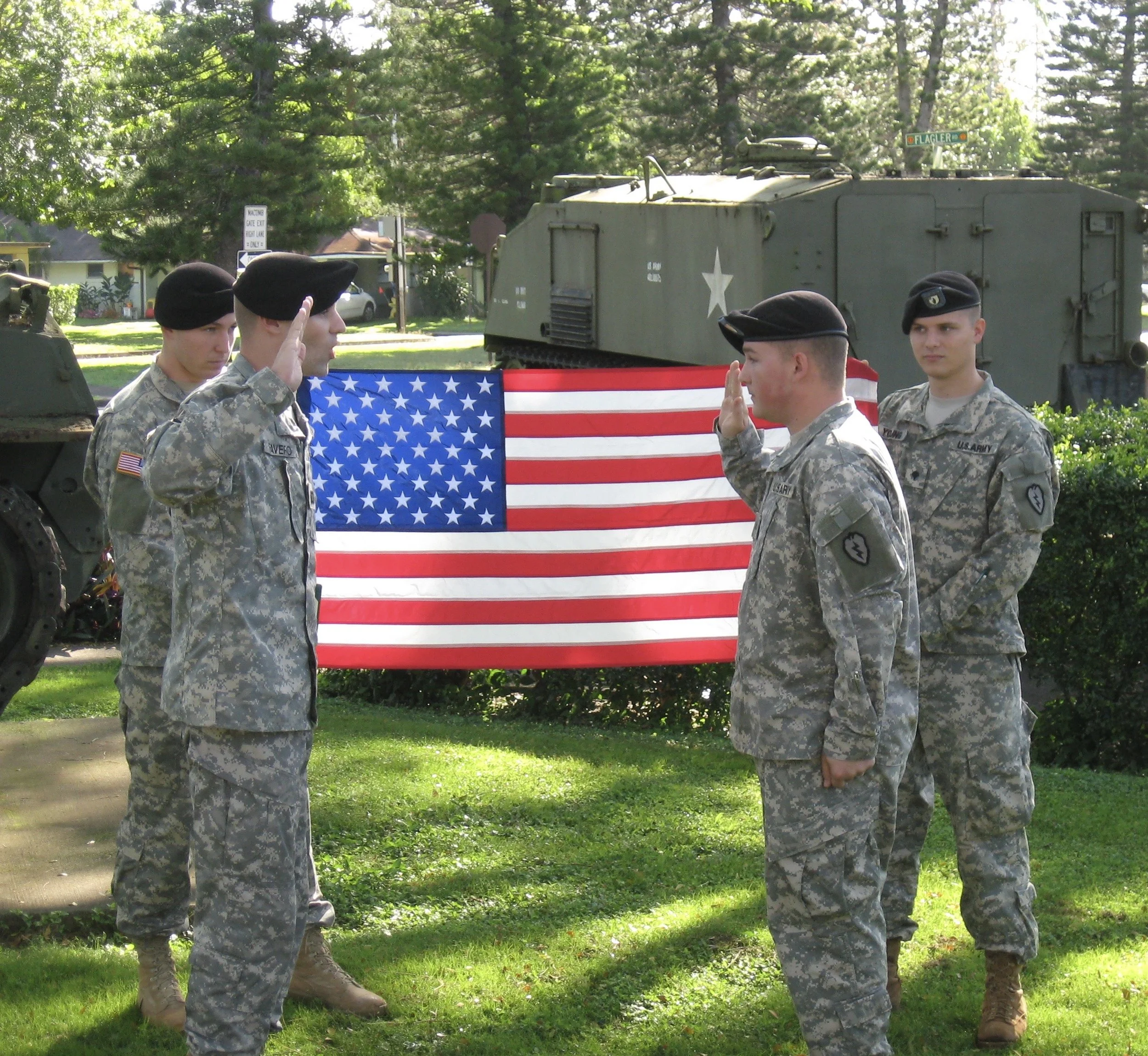 Four soldiers in camouflage uniforms and black berets standing in front of a military tank with a large American flag draped on the side. Two soldiers are saluting each other, and two others are watching. The scene is outdoors, with trees and houses in the background.