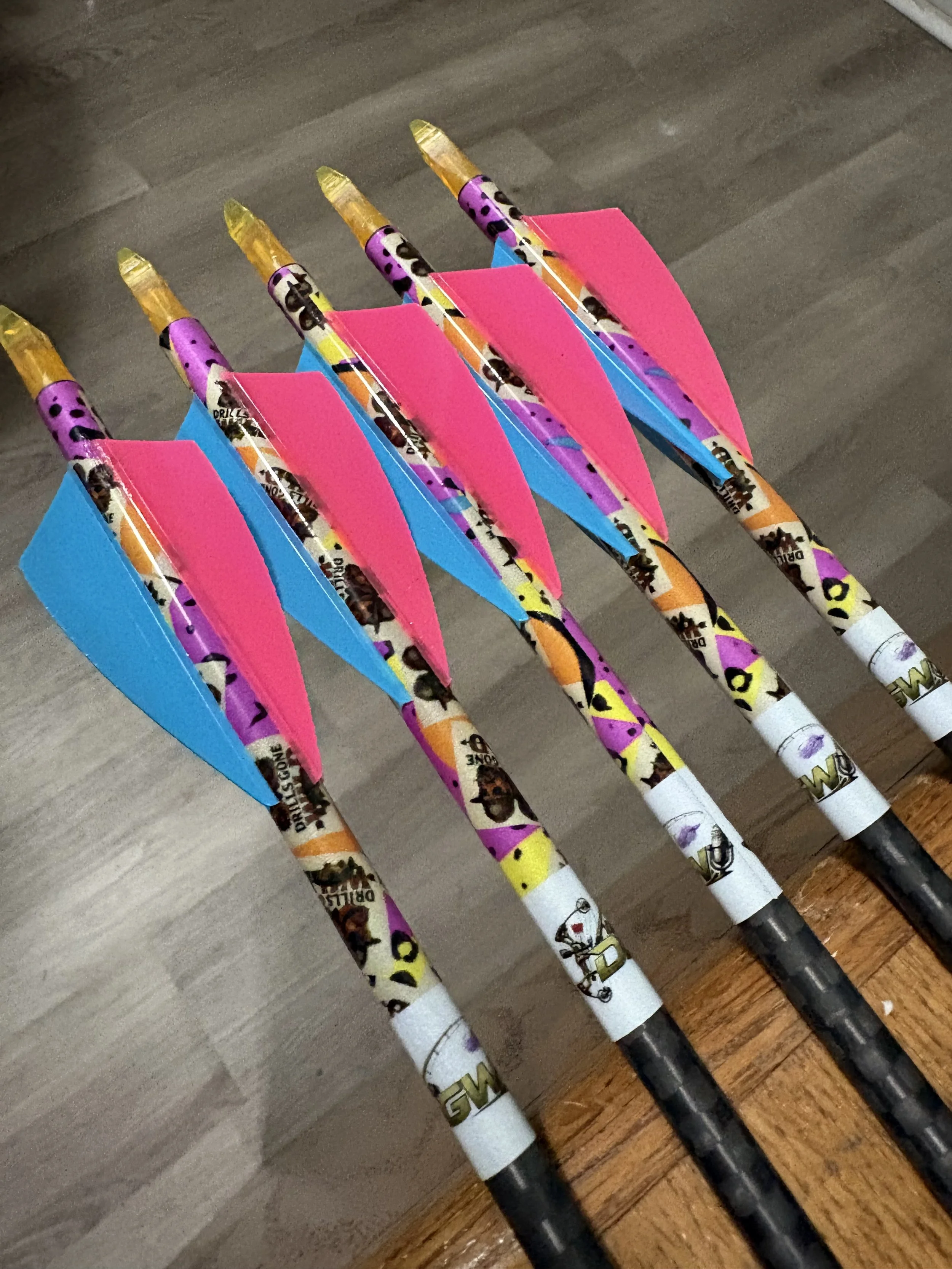 Six colorful arrows with pink, blue, and yellow decorations lined up on a wooden floor.