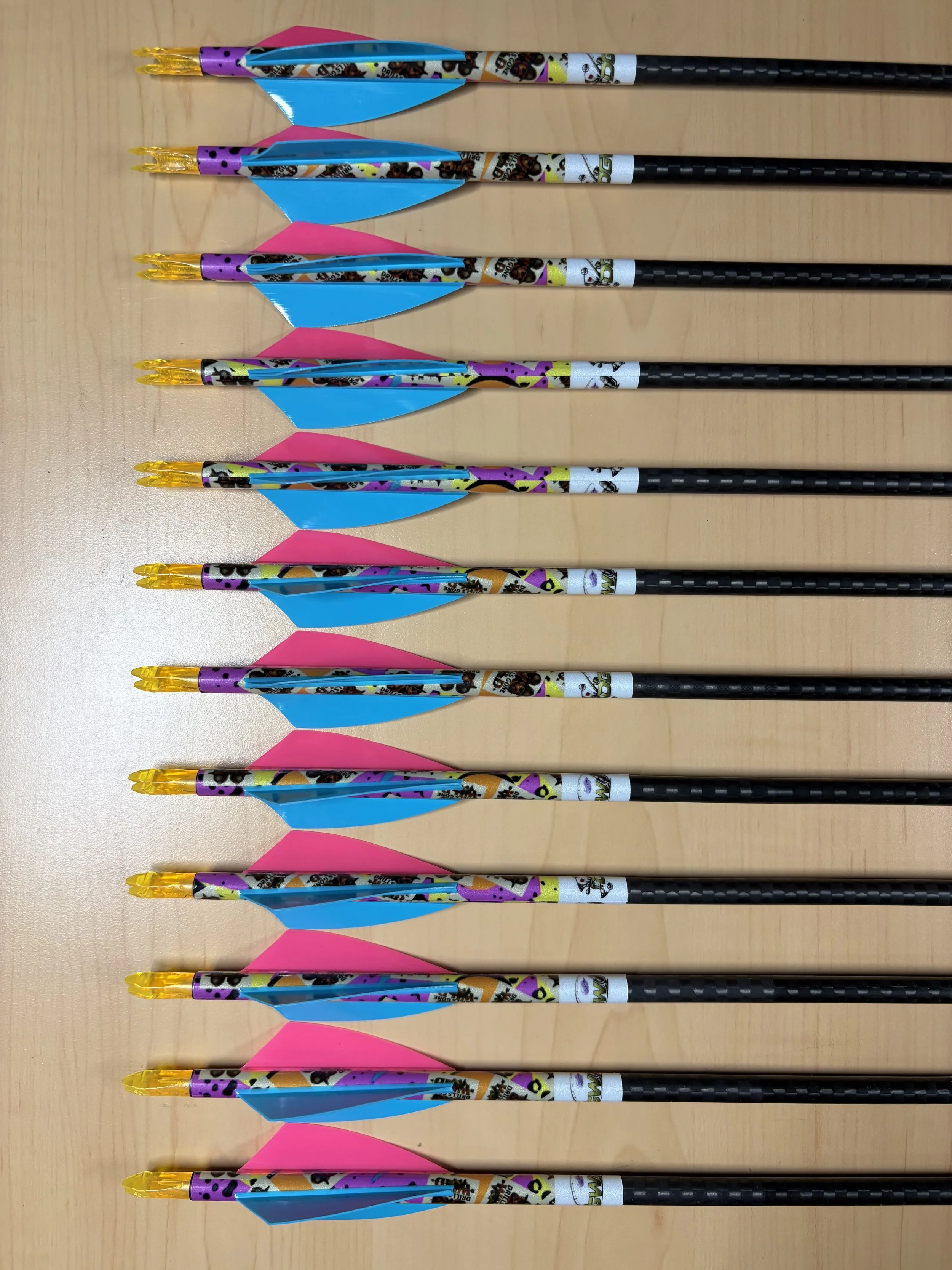 Six colorful arrows aligned horizontally on a wooden surface, with pink and blue fletchings, black shafts, and orange nocks.