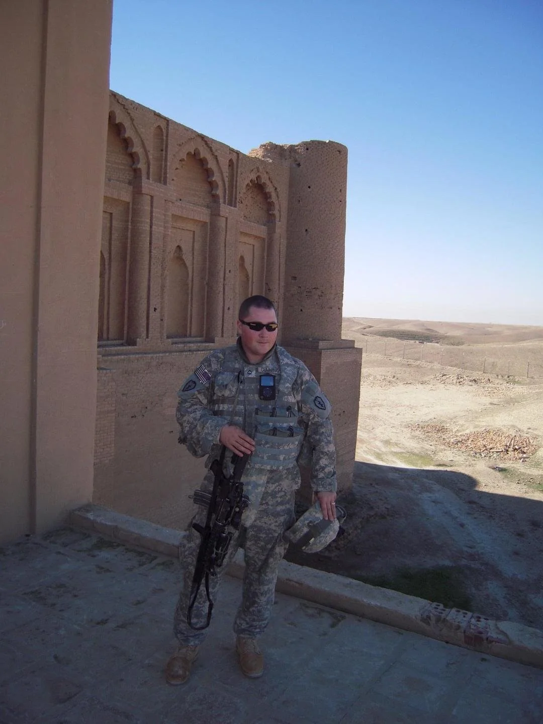 A soldier in camouflage uniform with sunglasses, holding a rifle, standing in front of an ancient, sand-colored brick building with arched windows and a tower in a desert landscape.
