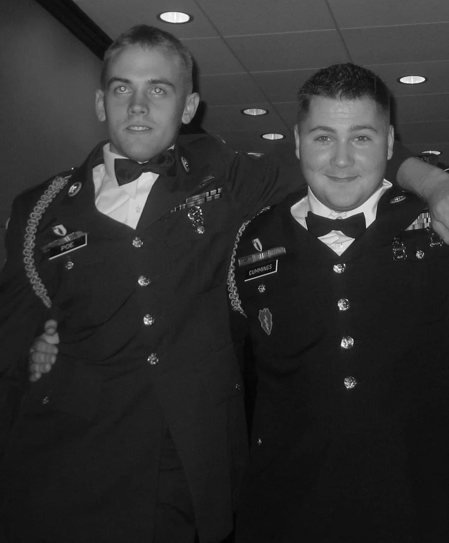 Two young men in formal military dress uniforms posing together with their arms around each other's shoulders, smiling at the camera, in an indoor setting with ceiling lights.