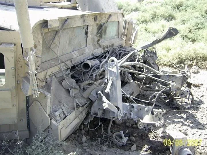 The image shows a heavily damaged, burned electrical vehicle or machinery parts in a desert-like environment, with wires and metal components scattered and fused together.