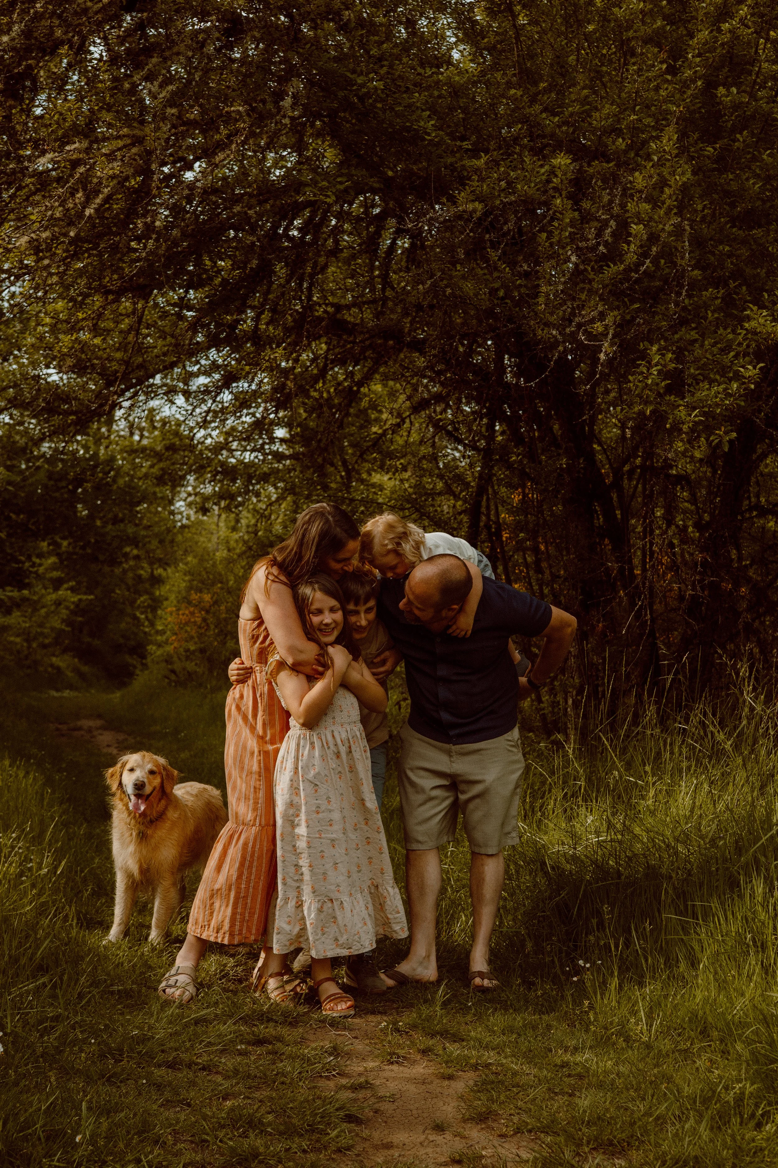 A family of five including children and adults, with a dog, standing outdoors in a wooded area during daytime, smiling and looking downward together.