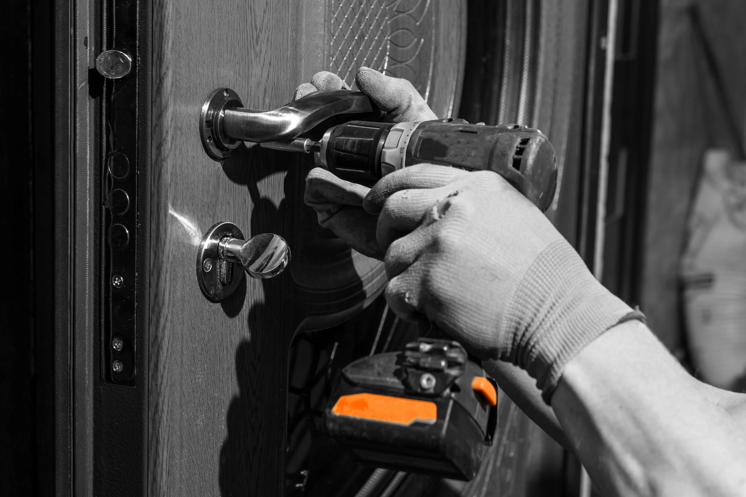 A person using a cordless drill to install a door handle on a wooden door. The person is wearing a glove on their left hand, and the door has a keyhole and a round doorknob.