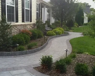 Curved stone walkway surrounded by landscaped plants and trees in front of a house with siding and large windows.
