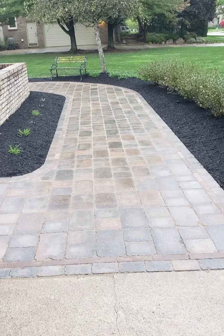 Brick paver walkway through landscaped garden area with bench, shrubs, trees, and residential hardscaping.