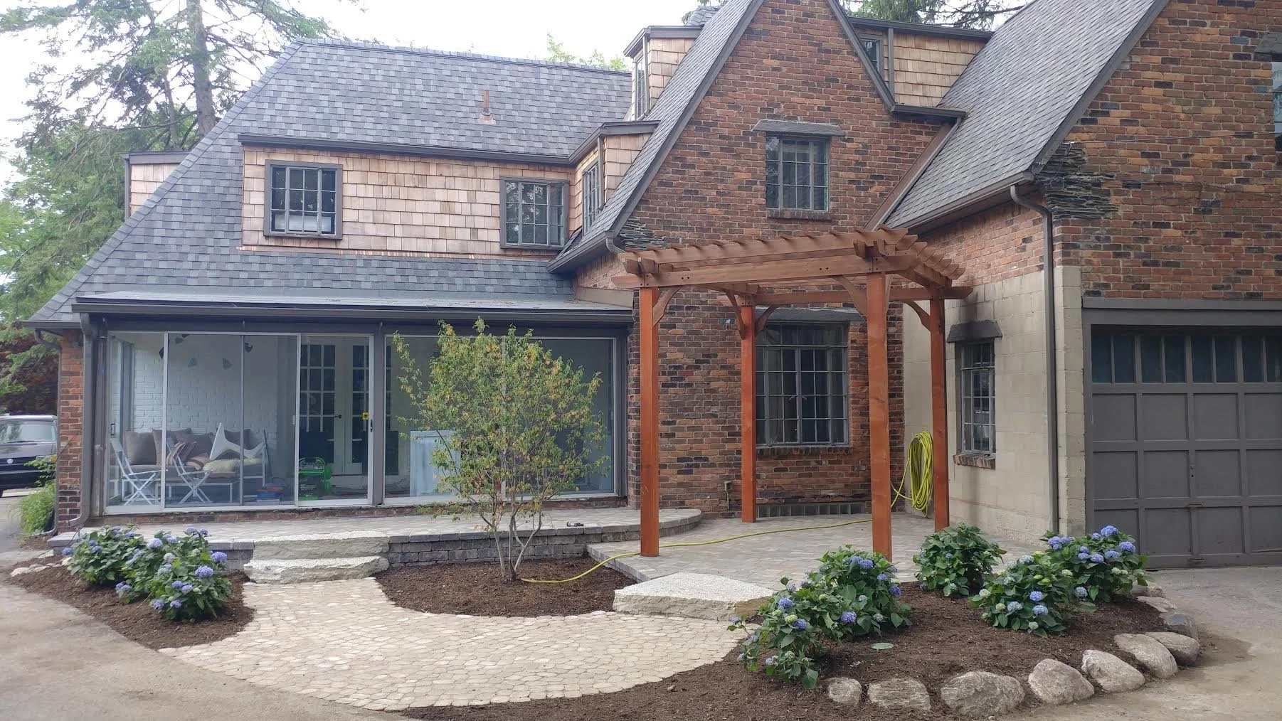 A entryway of a brick house, a small tree, and flower beds with purple hydrangeas, a stone walkway, and a wooden pergola.