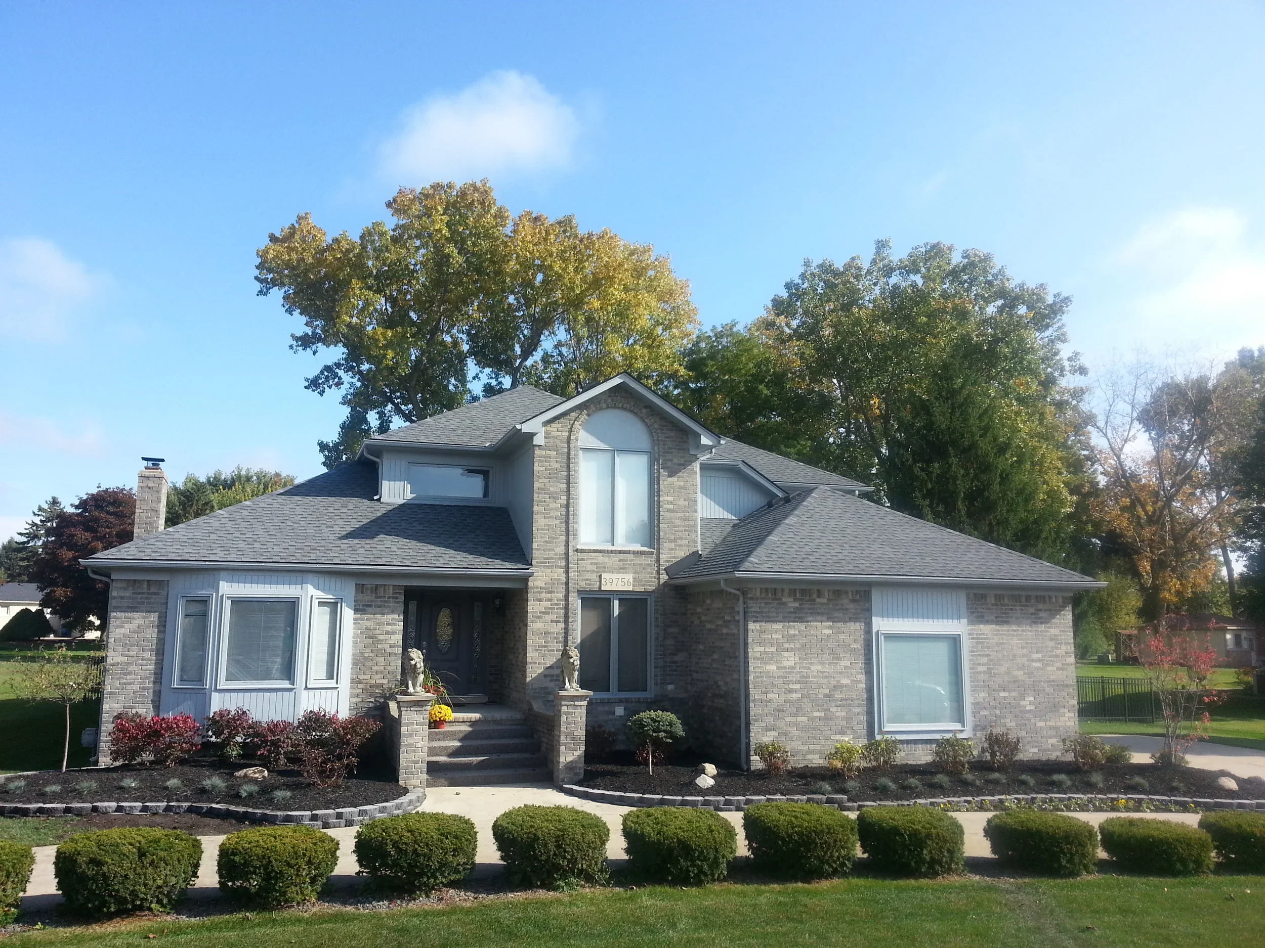 Professional front yard landscaping with trimmed lawn, shrubs, flower beds, mature trees, and decorative front porch features.
