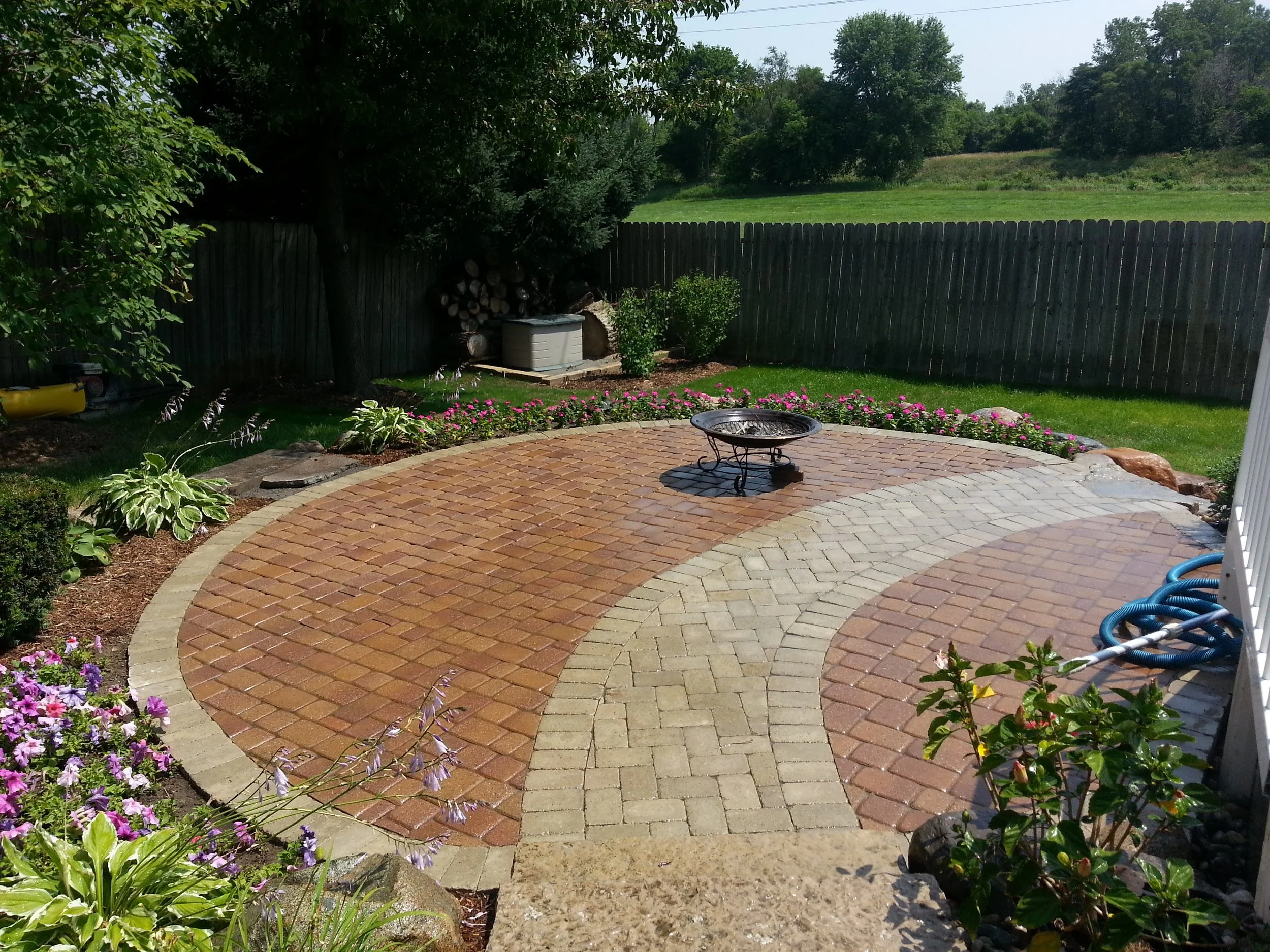Backyard paver patio with circular brick and stone design, flower beds, fire pit, and outdoor living area.