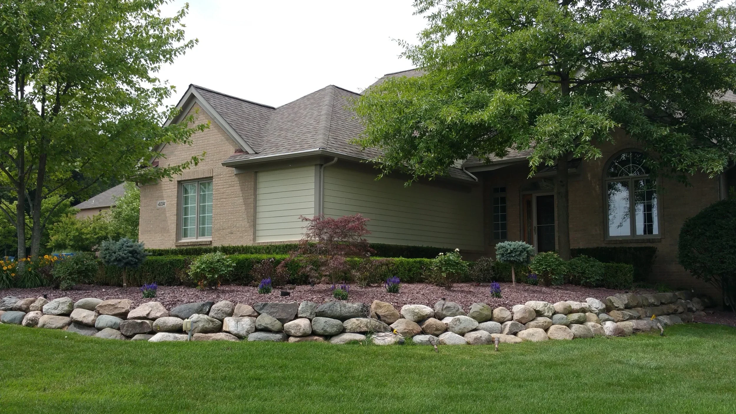 Residential landscaping with maintained lawn, shrubs, colorful flowers, trees, and stone garden bed border.