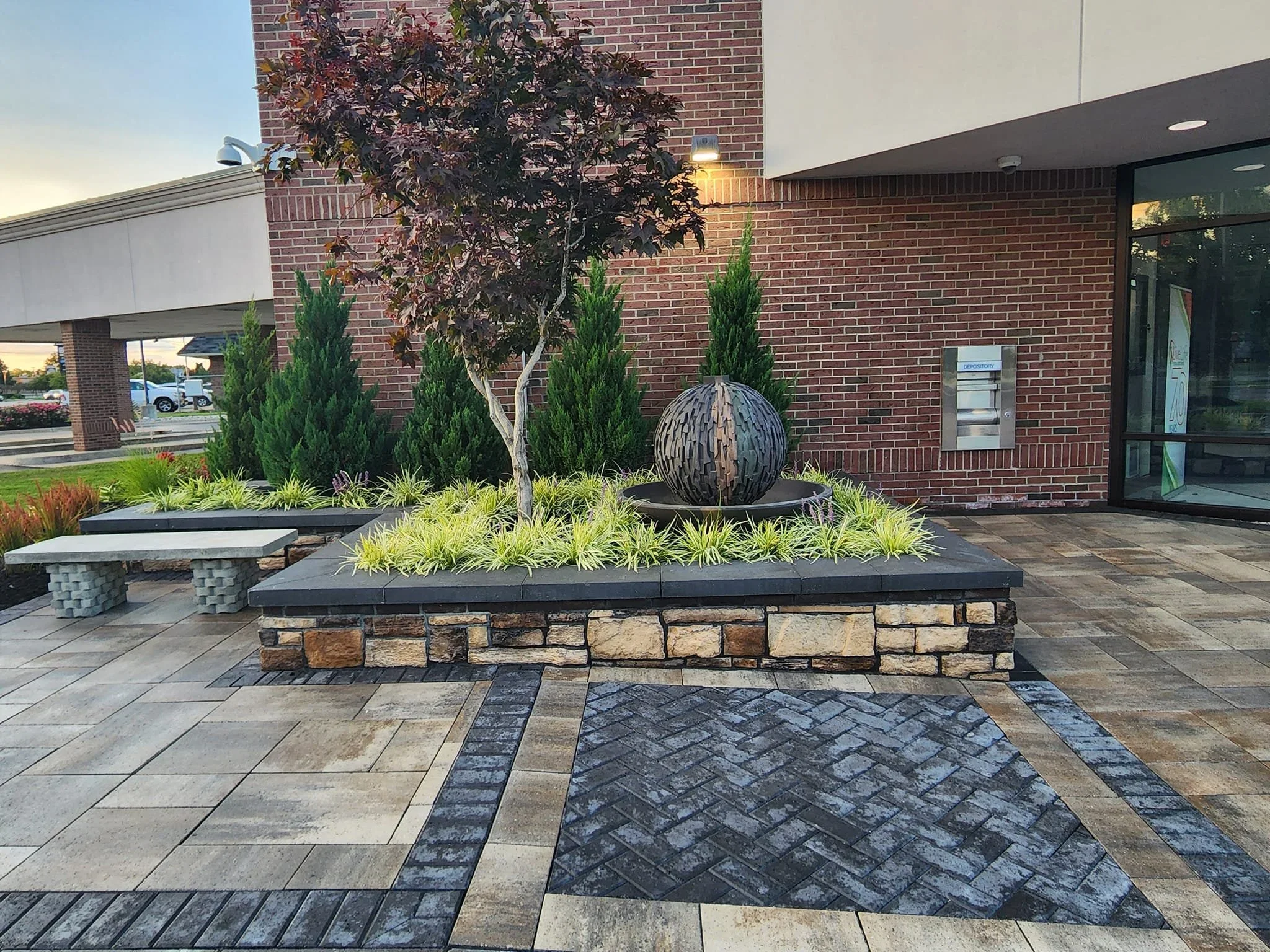 Commercial landscaping with decorative stonework, shrubs, small tree, seating area, and building entrance design.