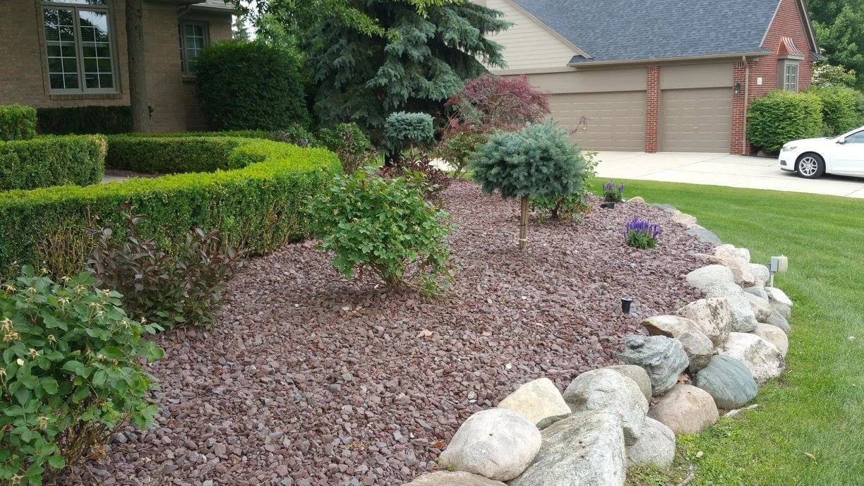 A landscaped residential front yard with neatly trimmed bushes, small trees, purple flowers, decorative rocks, and a well-maintained lawn. There's a house with brick and siding in the background, along with a driveway and a parked white car.