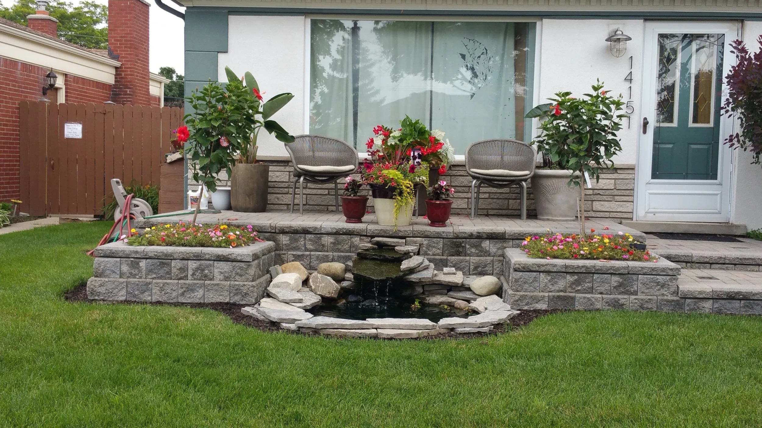 Front porch landscaping with potted plants, seating, rock border, and small decorative pond.