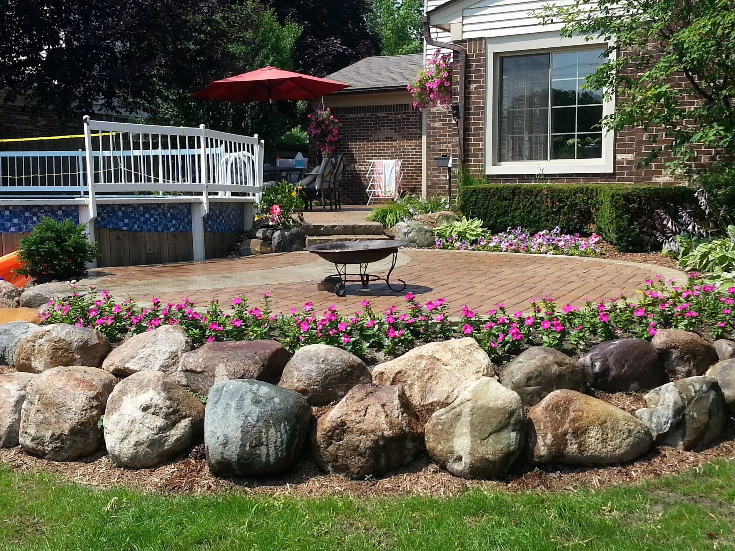 Backyard patio landscaping with rock garden border, flower beds, outdoor seating, umbrella, and hot tub area.