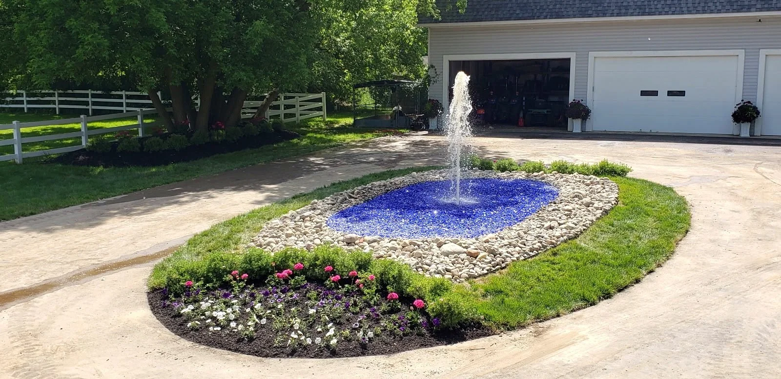 Custom driveway island water feature with fountain jet, blue glass, river rock border and flower bed