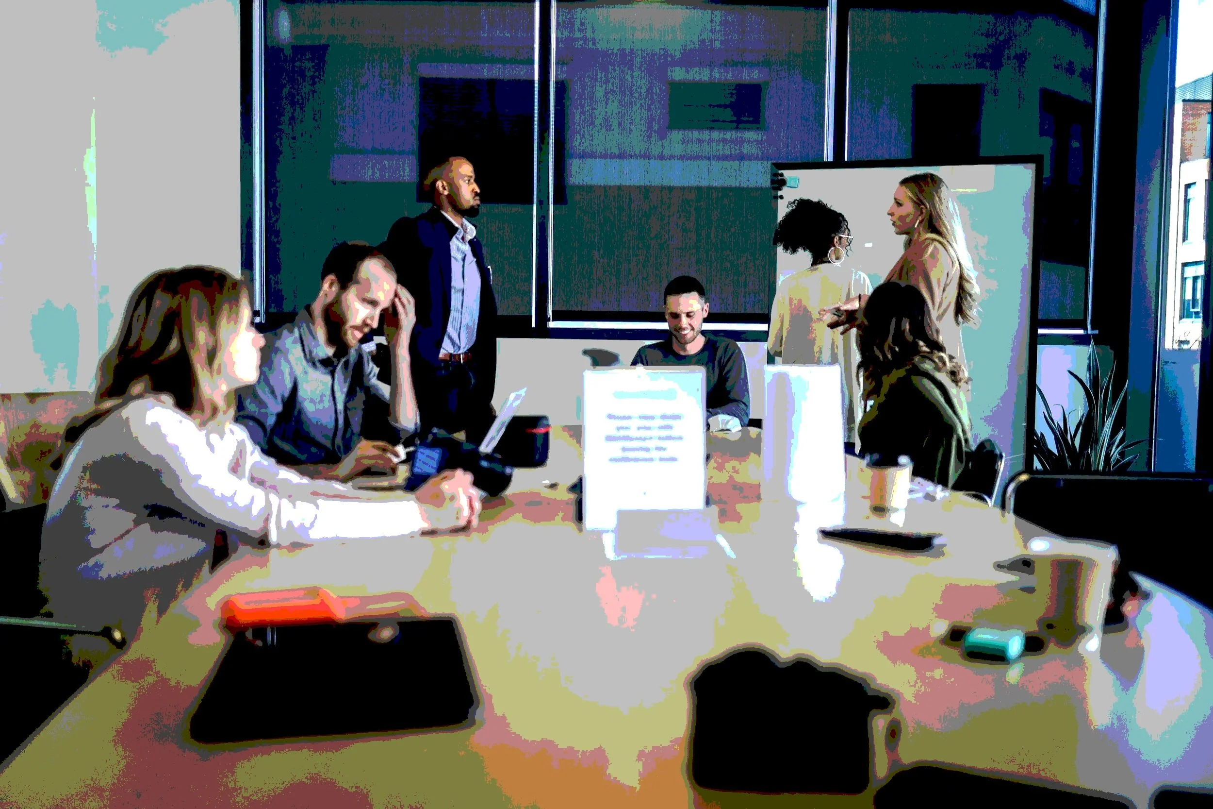 People in an office meeting, some seated with laptops and others standing and talking