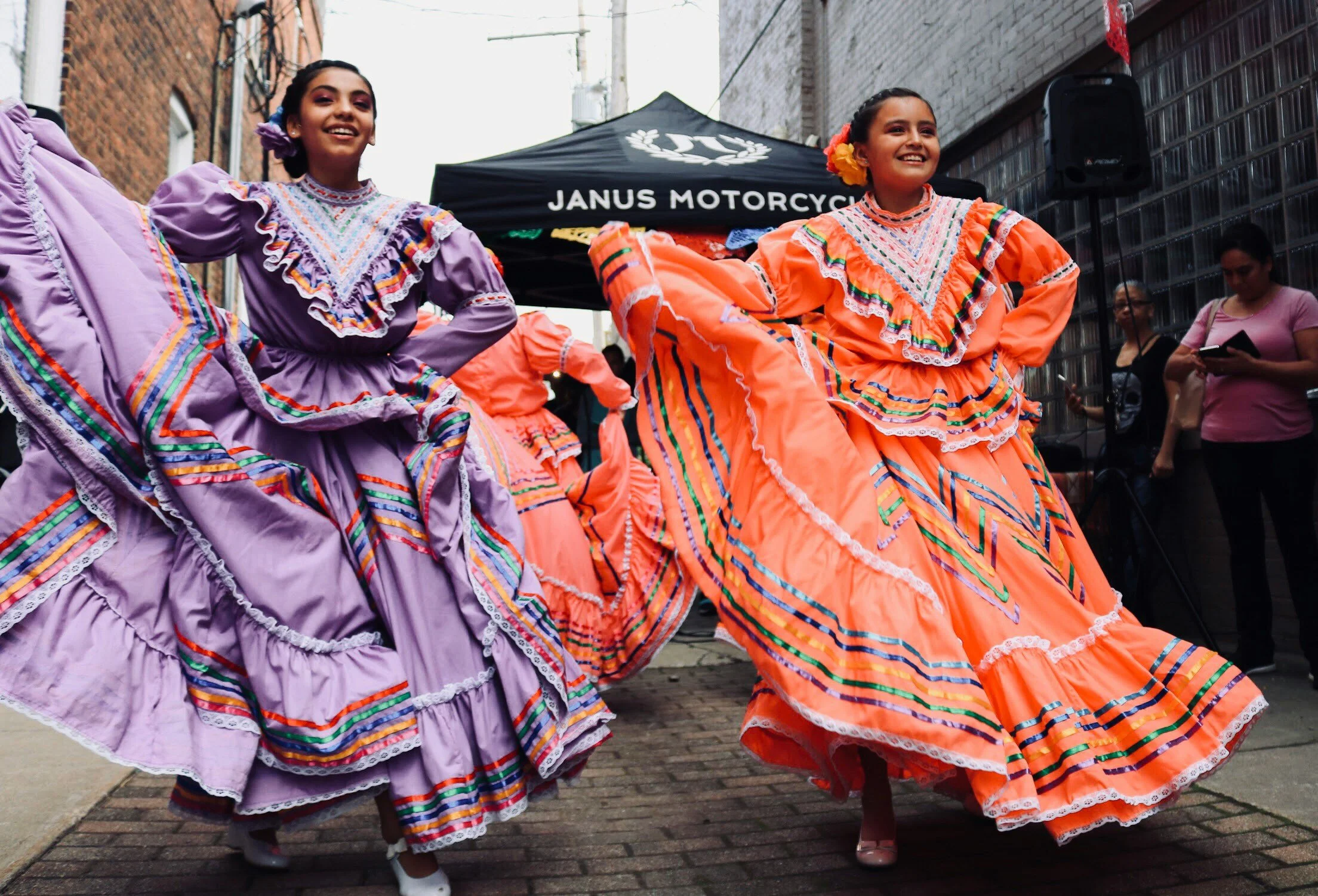 Two women wearing colorful traditional Mexican dresses, dancing on a street during a celebration or cultural event.