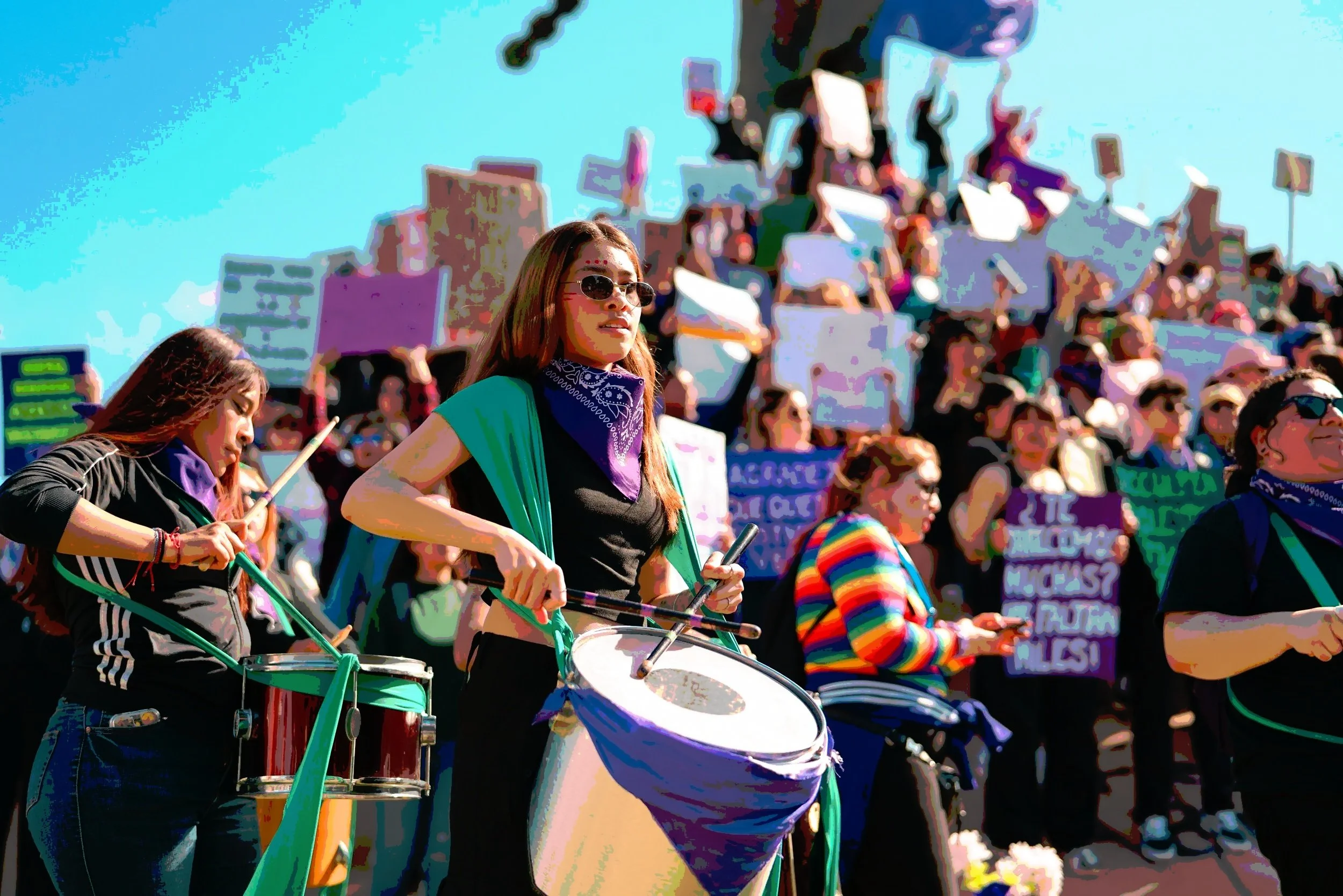 Women playing drums and holding signs in Spanish at a community protest, illustrating grassroots resistance