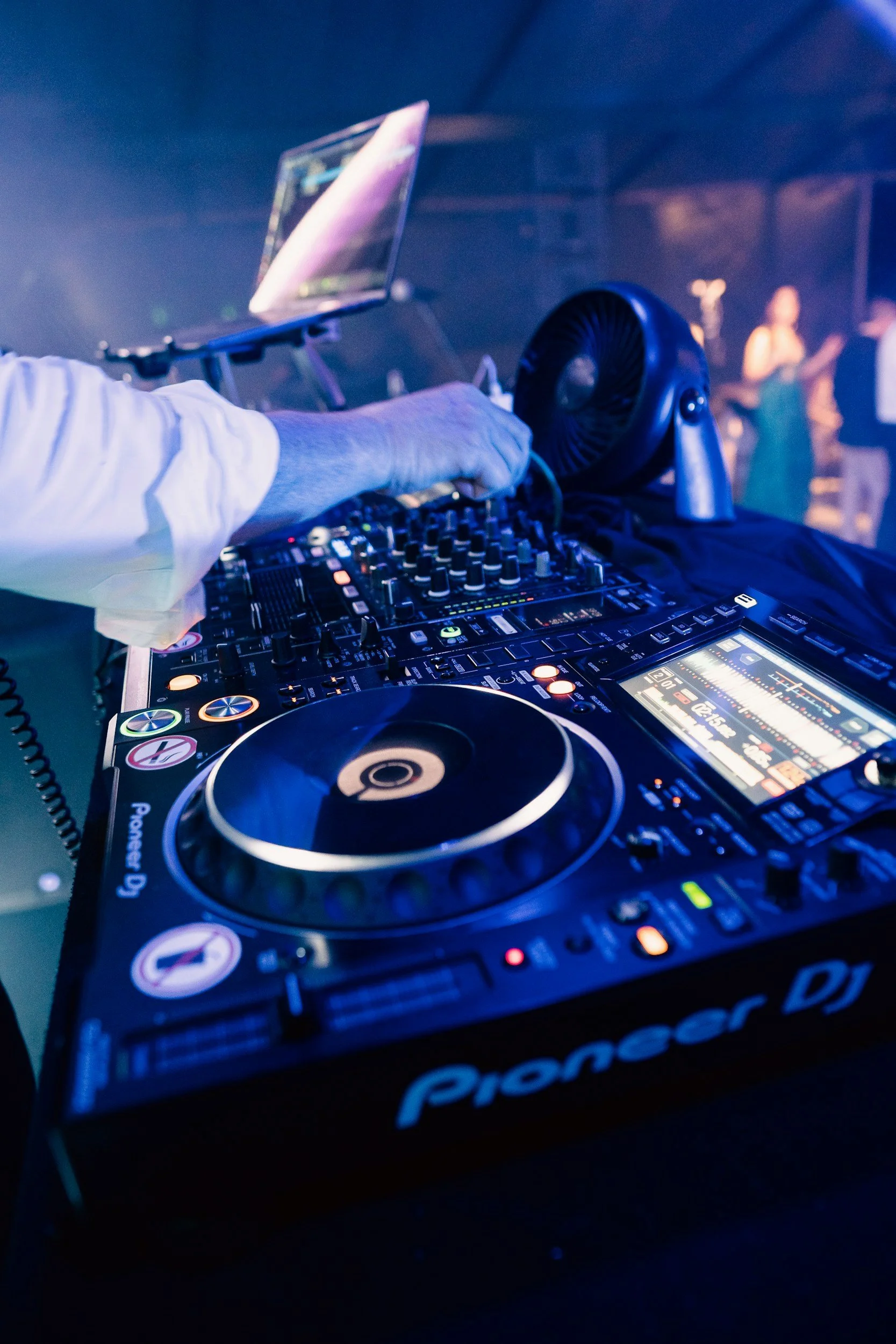 Close-up of DJ equipment, including a Pioneer DJ controller and a laptop on a stand, with a blurred background of people dancing in a club or party setting, illuminated by colorful lights.