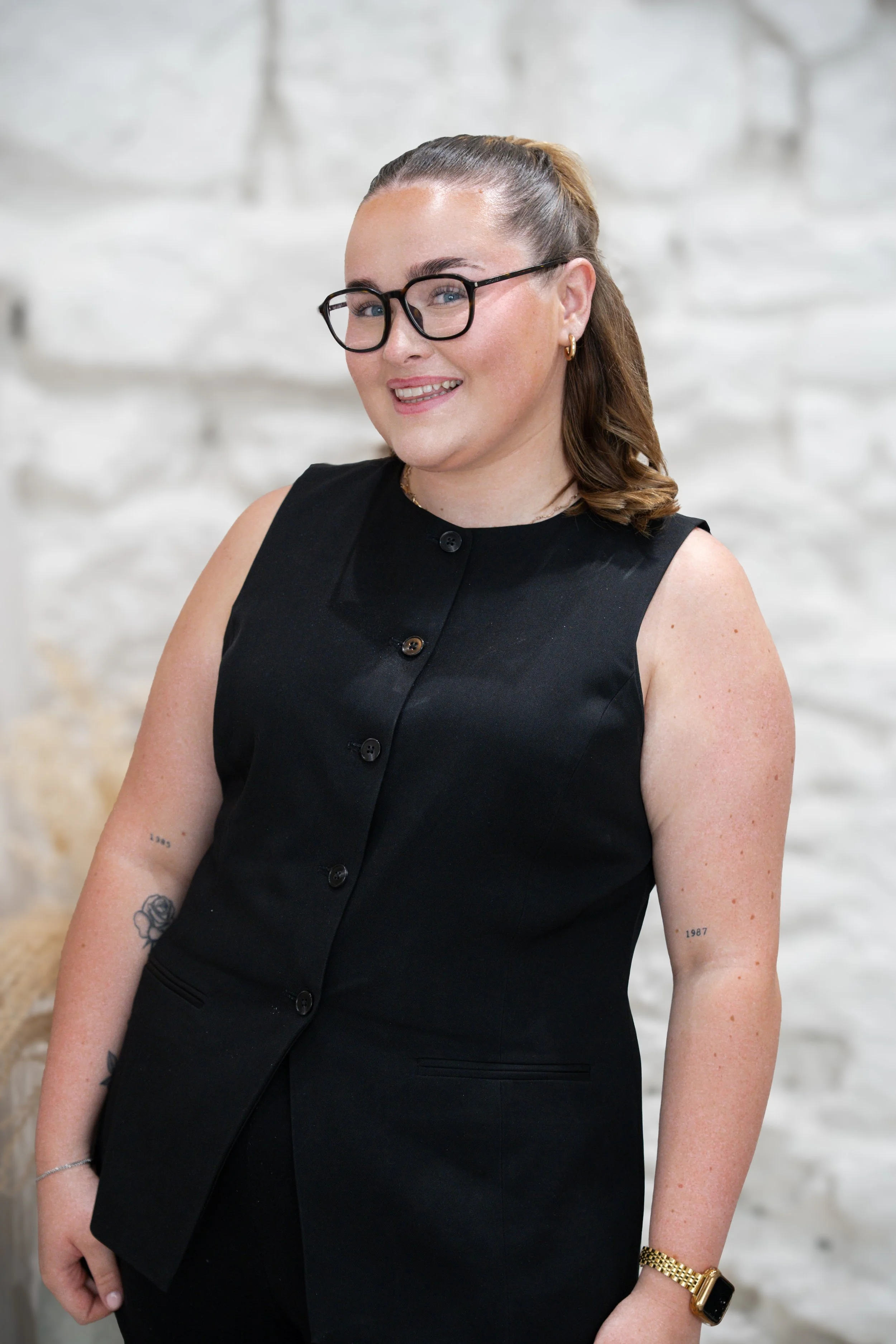 A woman with long brown hair tied in a ponytail, wearing black glasses, a black sleeveless button-up shirt, earrings, a gold watch, and tattoos on her arms, standing in front of a white textured wall.