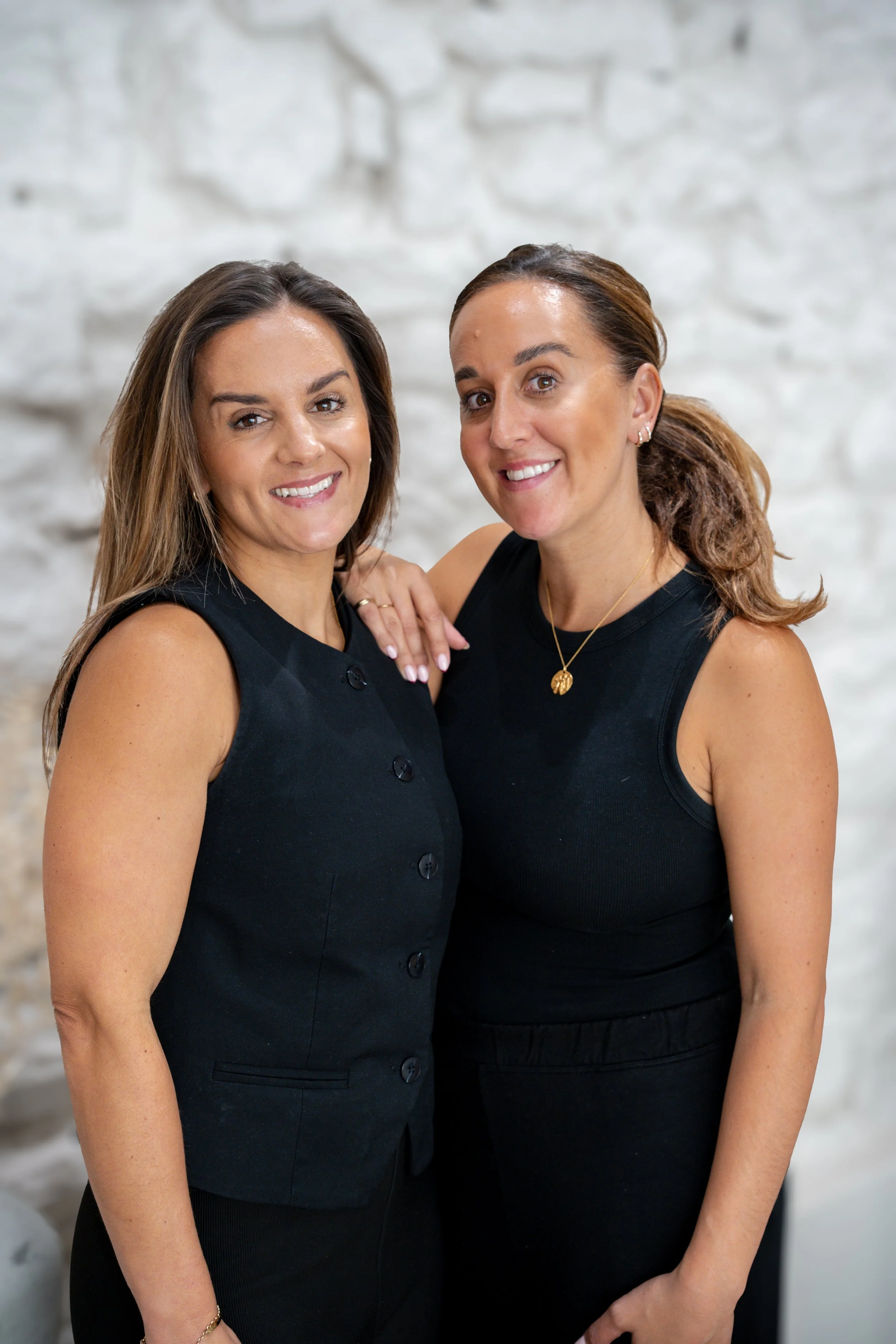 Two women standing closely together, smiling, against a white brick wall, both wearing black sleeveless tops.