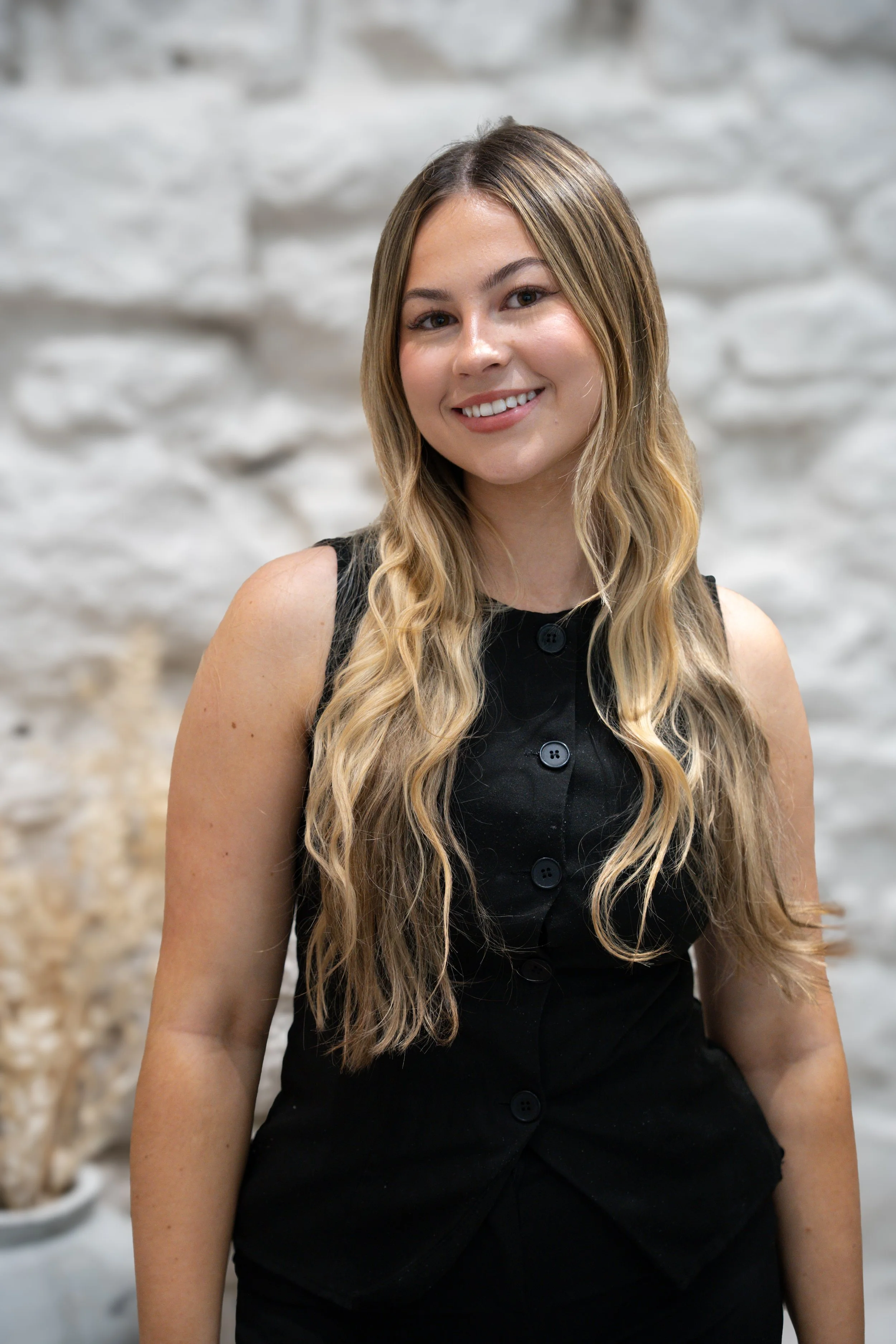 A young woman with long wavy blonde hair, wearing a sleeveless black dress with buttons, smiling at the camera against a blurred white brick wall background.