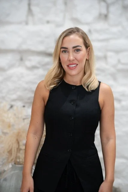 A woman with blonde hair smiling, wearing a sleeveless black dress, standing in front of a white brick wall.