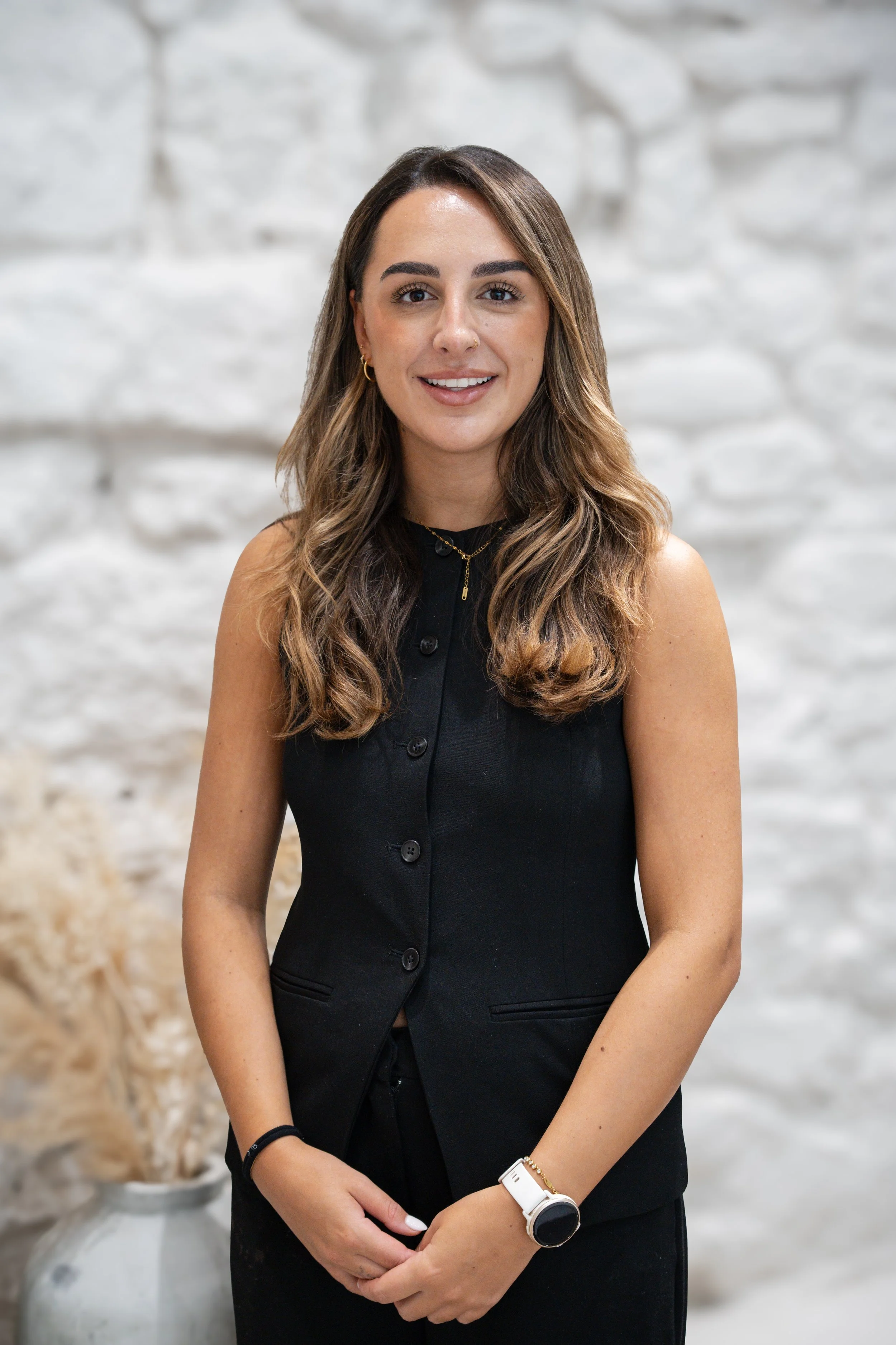 A young woman with long wavy hair wearing a sleeveless black button-up top, gold jewelry, and a white smartwatch posing in front of a white brick wall.