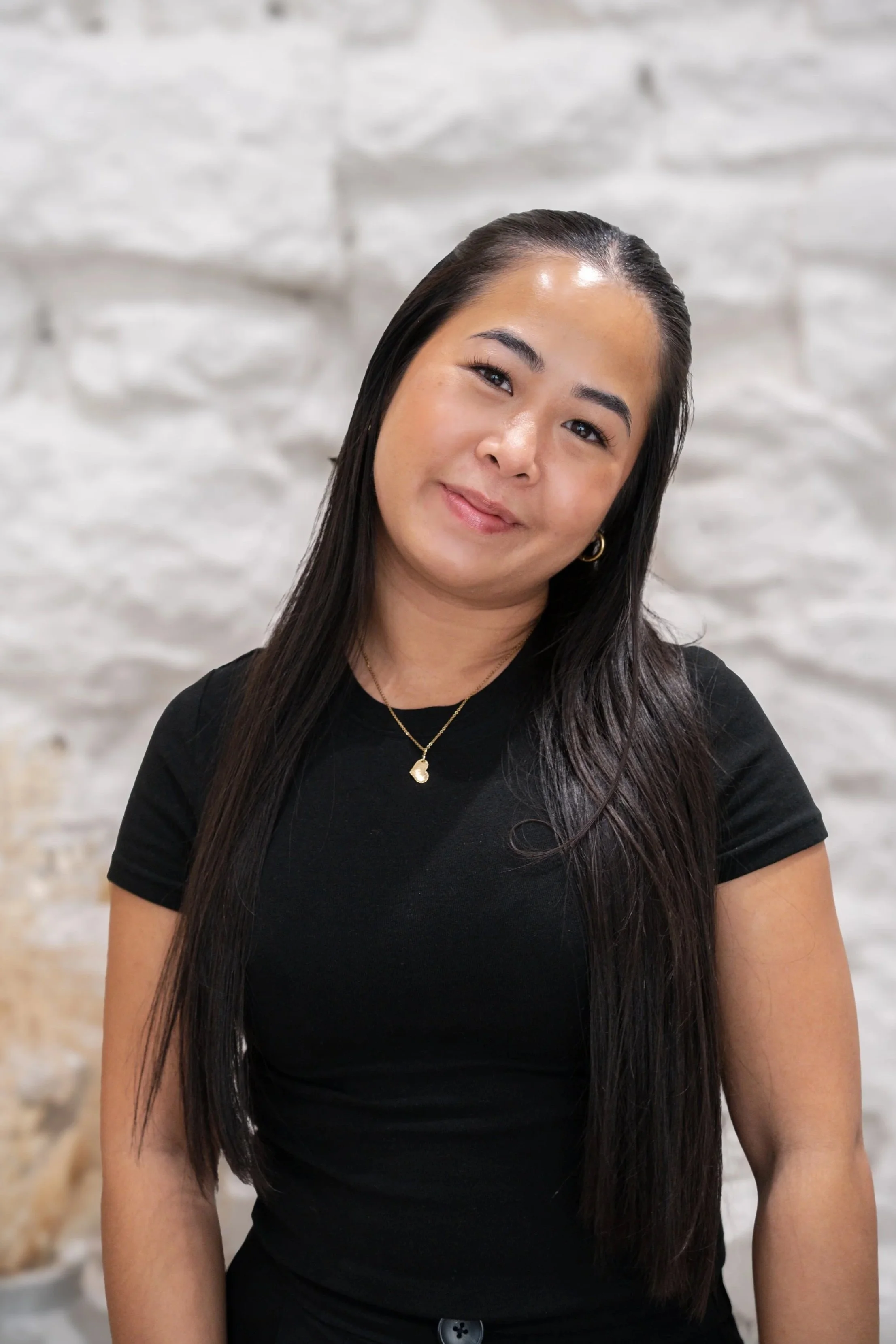 Portrait of a woman with long dark hair, wearing a black top, gold jewelry, and smiling softly, against a white brick wall background.