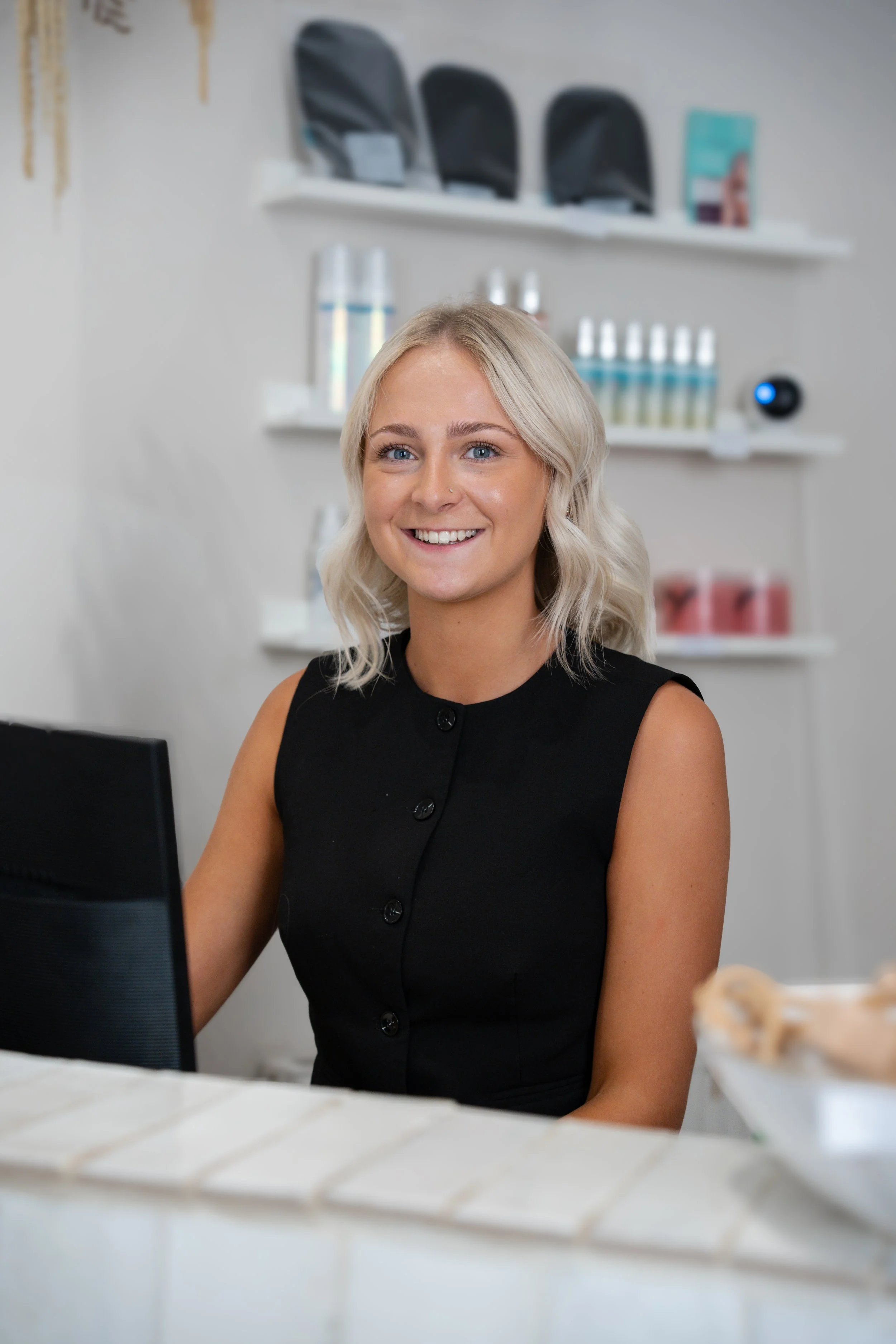 Smiling woman with blonde hair sitting at a desk in an office environment, with shelves displaying black bags, bottles, and a digital device in the background.