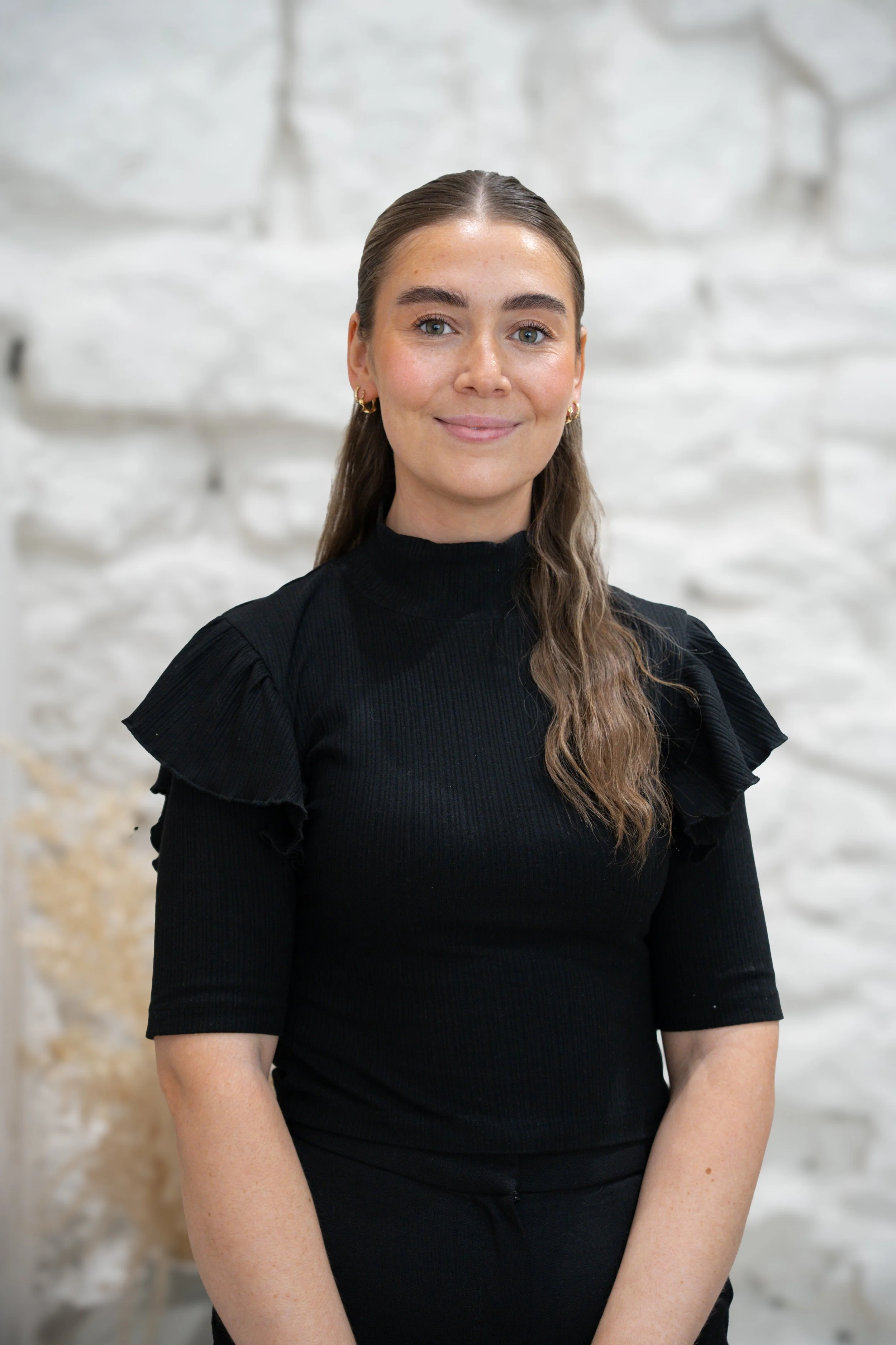 A woman with long brown hair, light skin, and wearing a black top standing in front of a white brick wall.