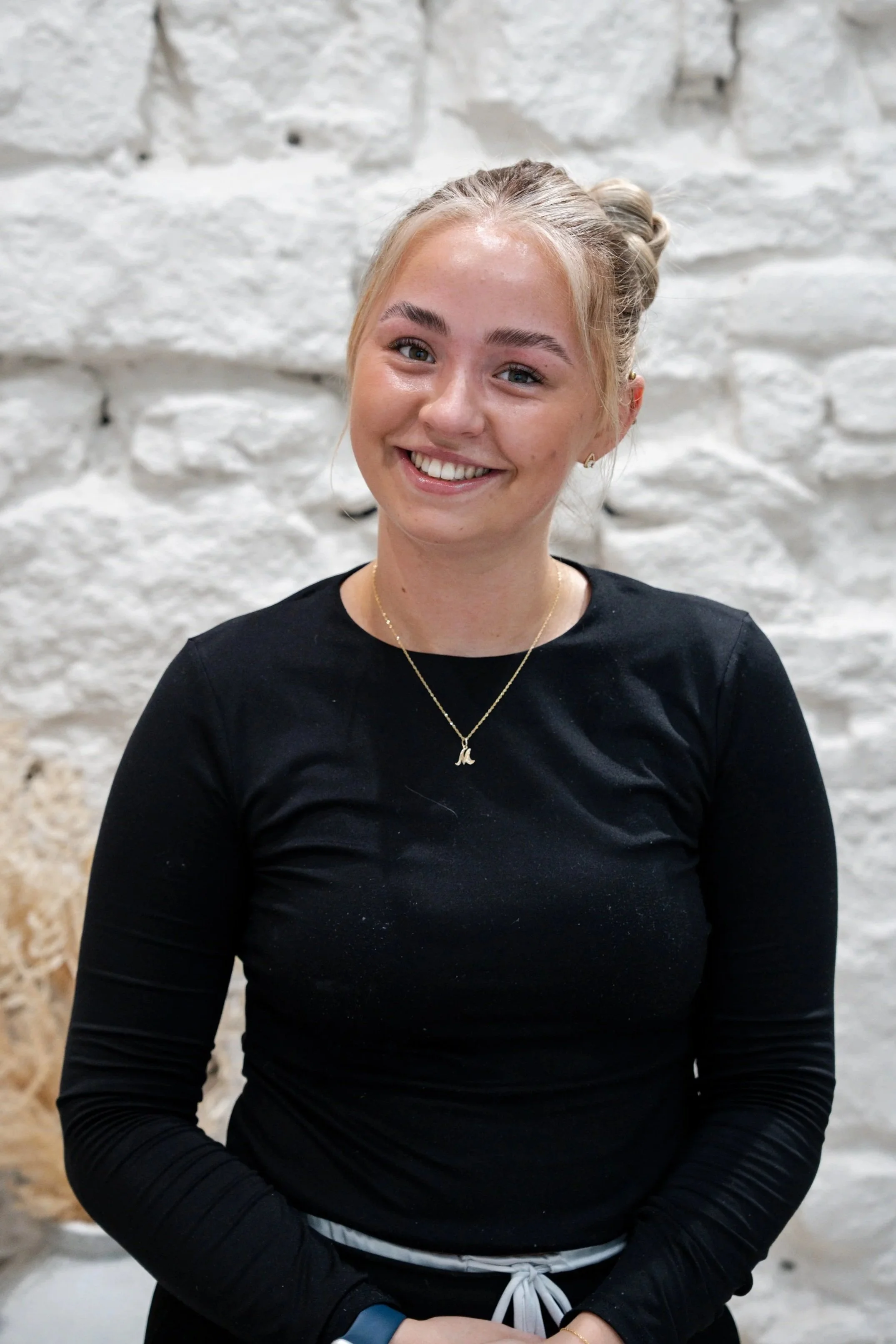Young woman with blonde hair tied in a bun, smiling, wearing a black long-sleeve top, gold necklace, earrings, and a bracelet, standing in front of a white brick wall.