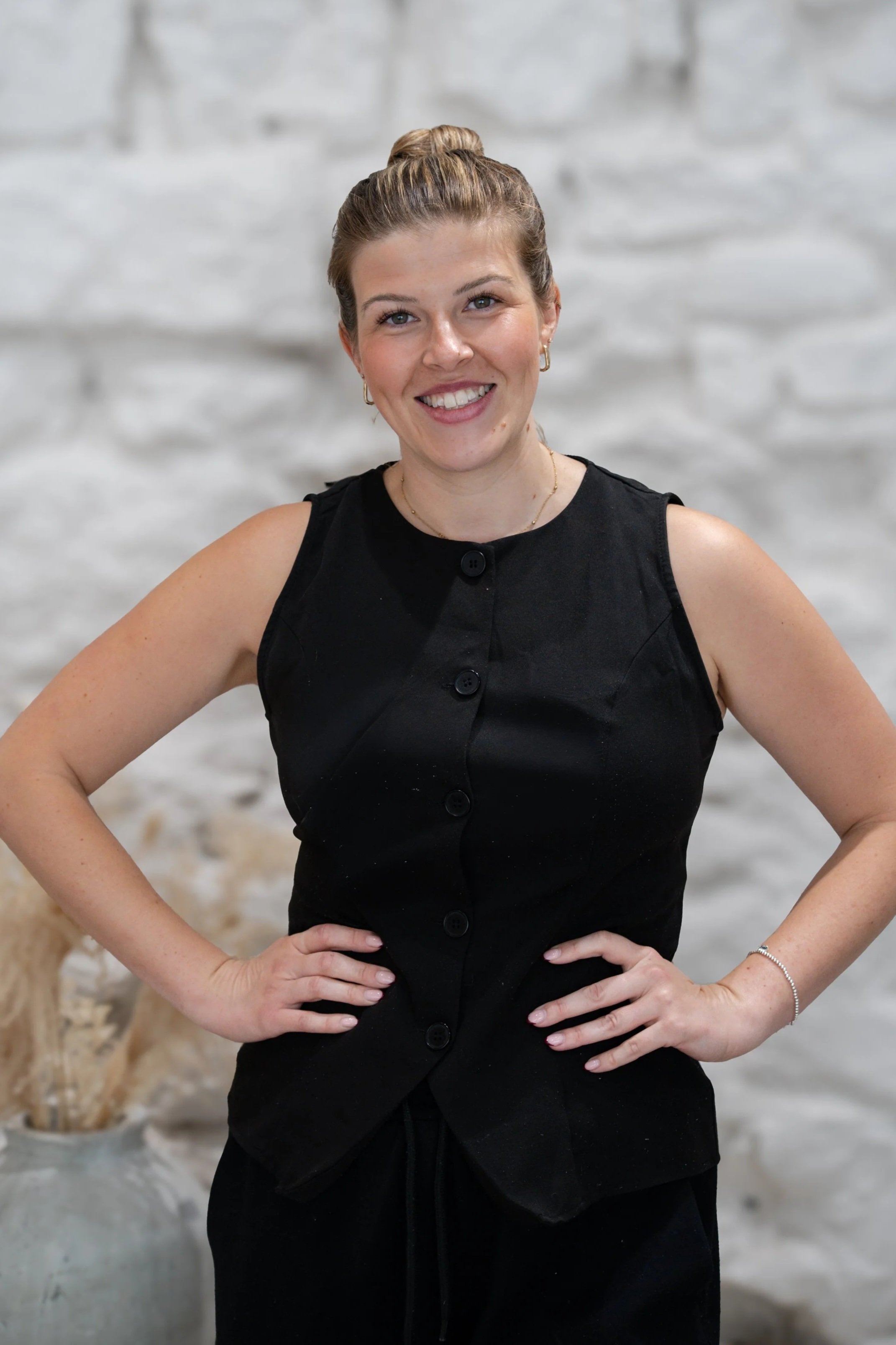 A woman with blonde hair in a bun wearing a sleeveless black top, smiling and standing with her hands on her hips. She is in front of a blurred white brick wall background.