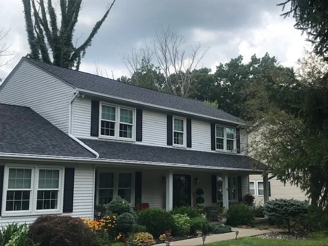 A two-story white house with black shutters, a front porch, and a well-maintained garden with various shrubs and flowers. Overcast sky with bare trees in the background.