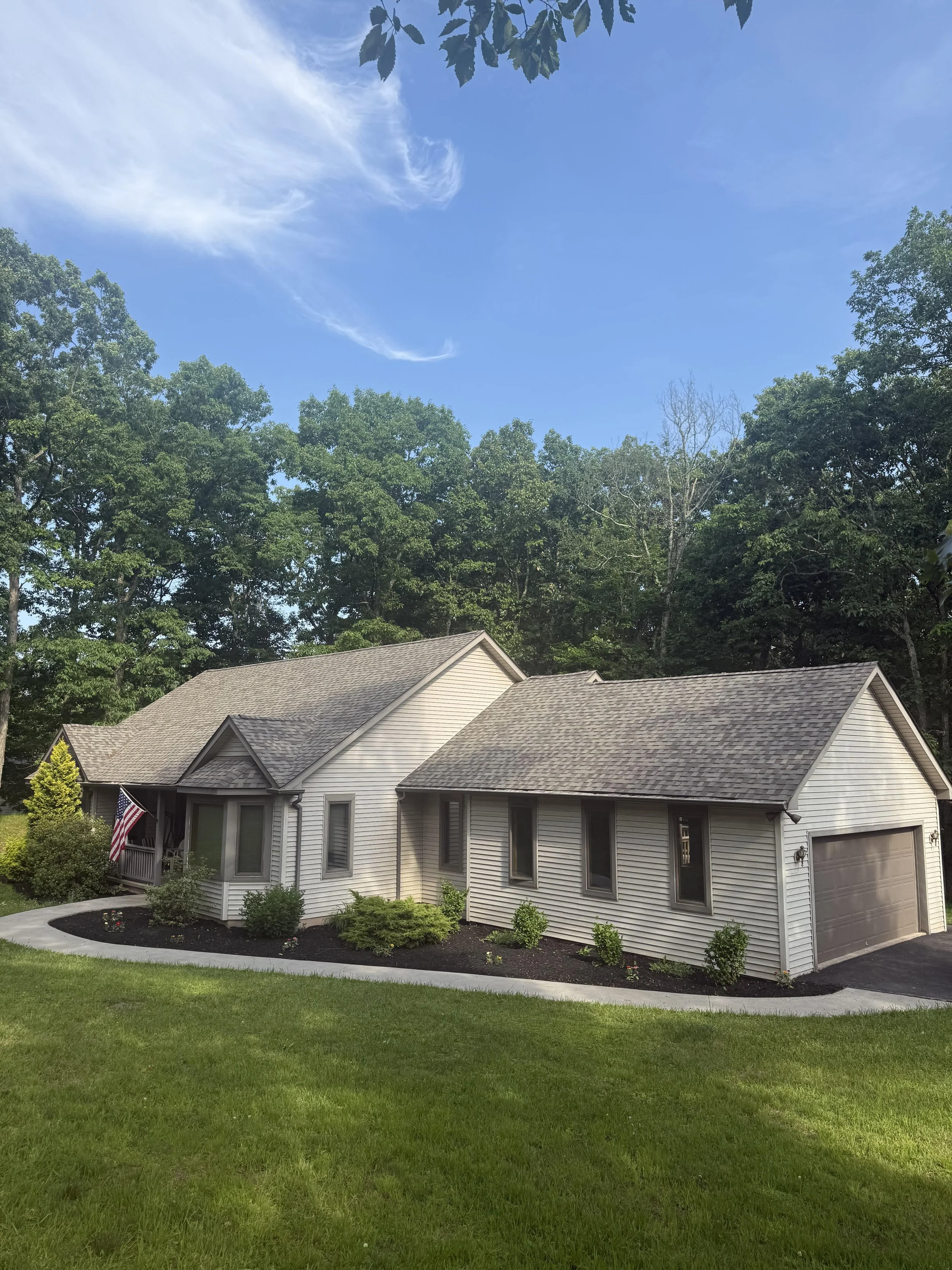 A suburban house with beige siding, a brown shingle roof, and multiple windows, surrounded by a well-maintained lawn, small bushes, and flowers. An American flag is displayed near the entrance. Tall trees and a blue sky with some clouds are in the ba