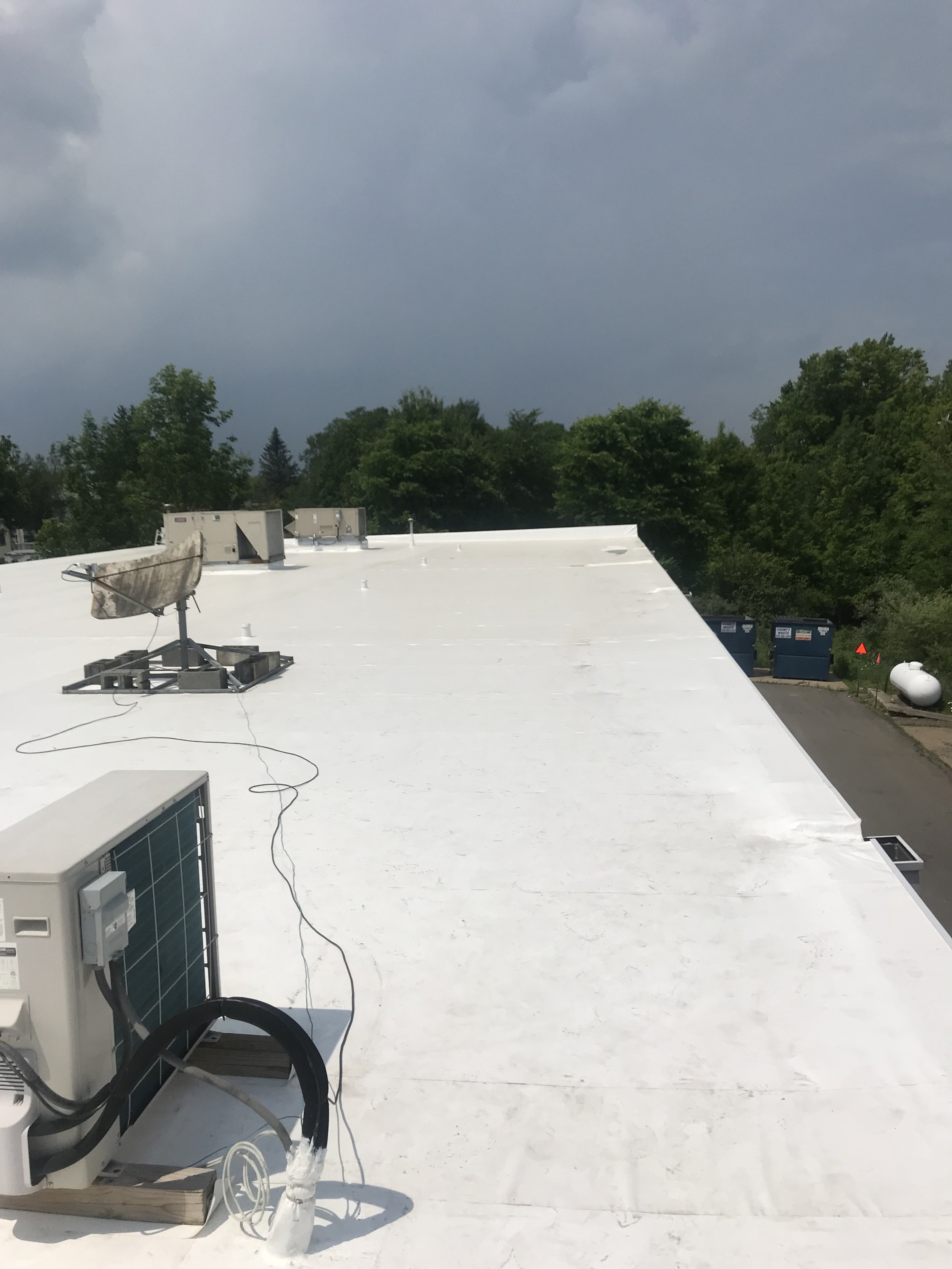 View of a white flat rooftop with some equipment including a satellite dish and an electrical box, overlooking trees and dark storm clouds in the sky.