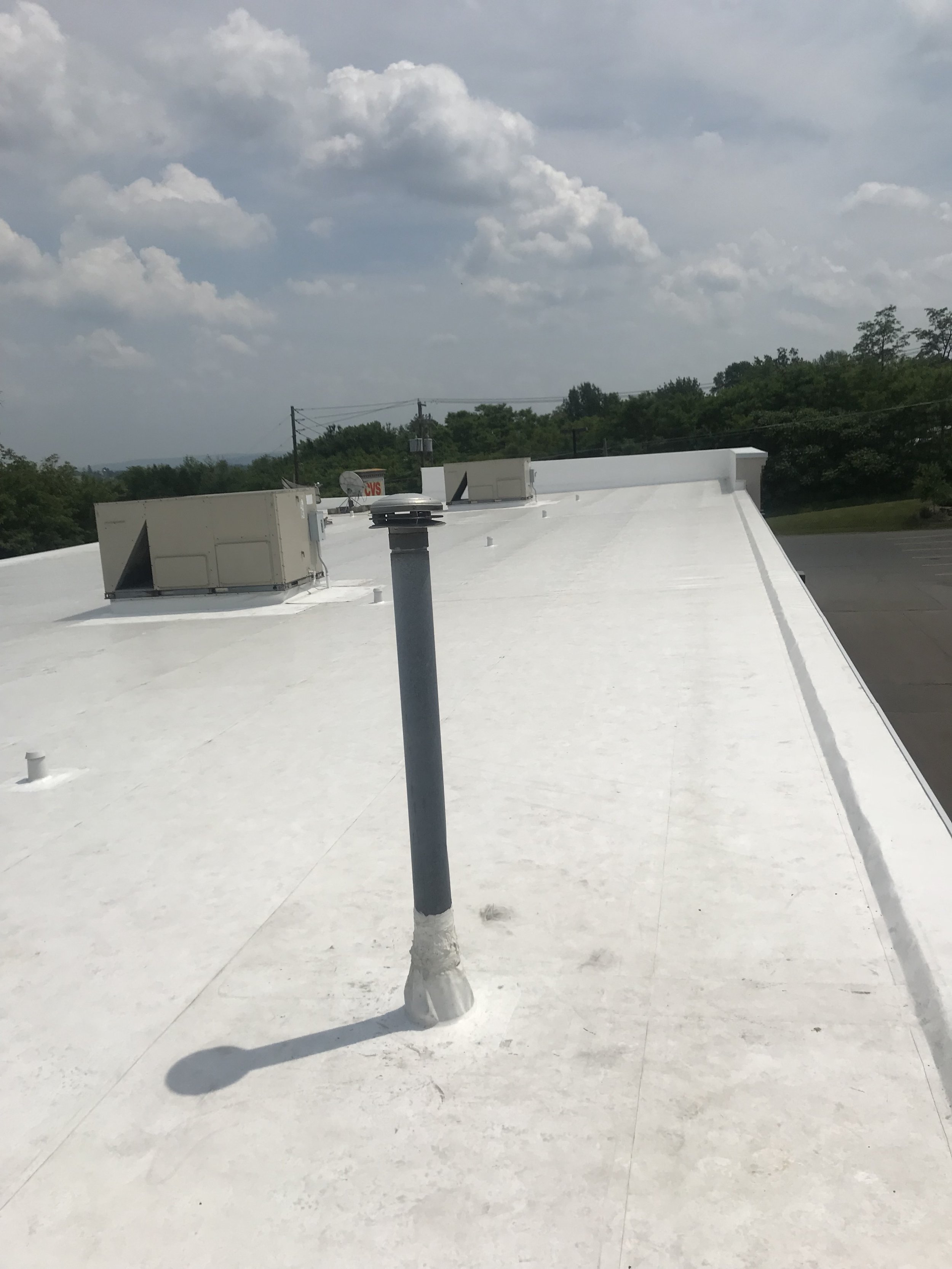 White flat roof with HVAC units, vents, and a pole casting a shadow. Background has trees, cloudy sky, and distant utility poles.