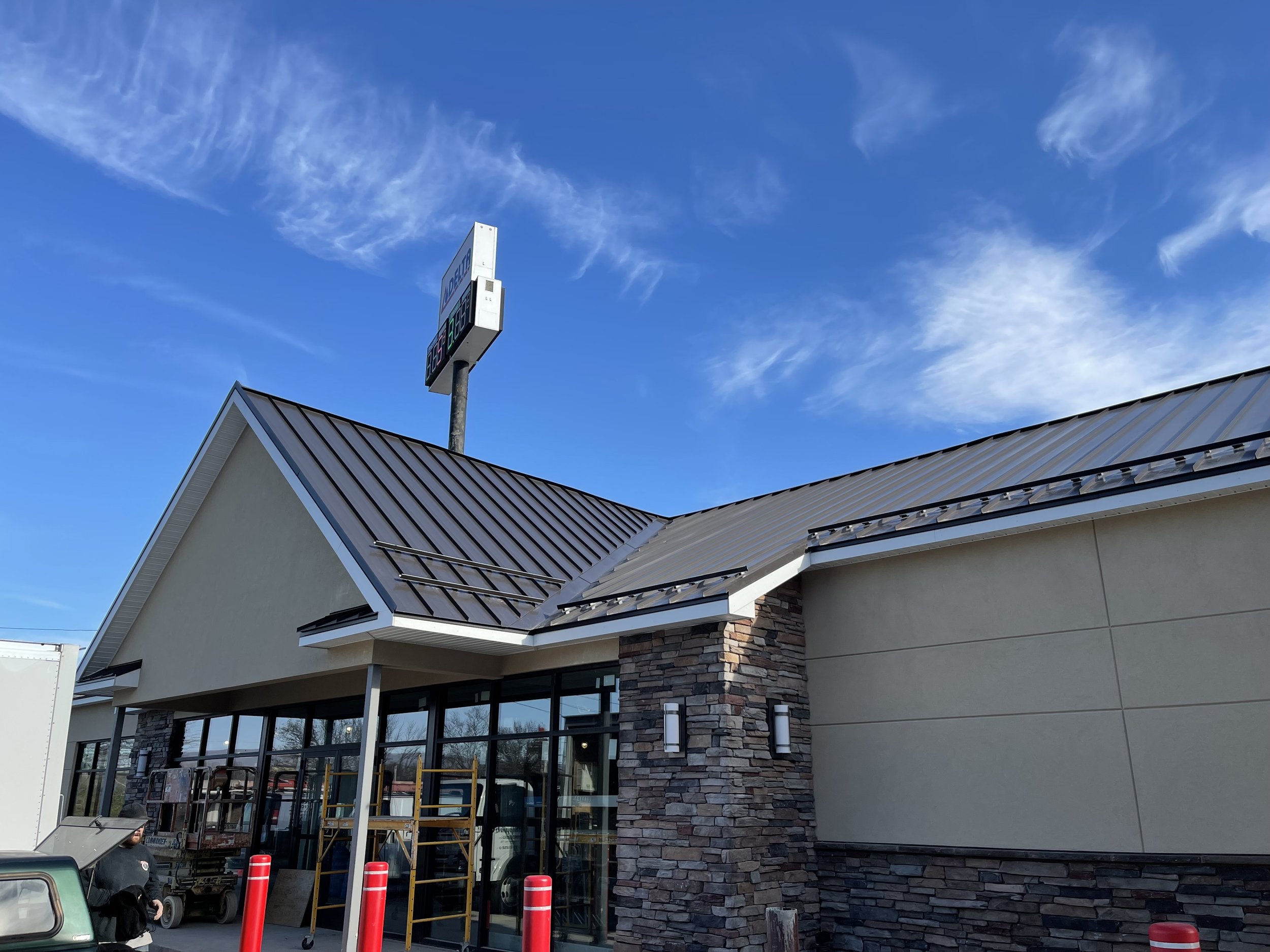 Front of a modern commercial building with a stone facade, large glass windows, and a metal roof. There is a sign on a pole in the parking lot, and orange traffic cones are visible near the entrance.