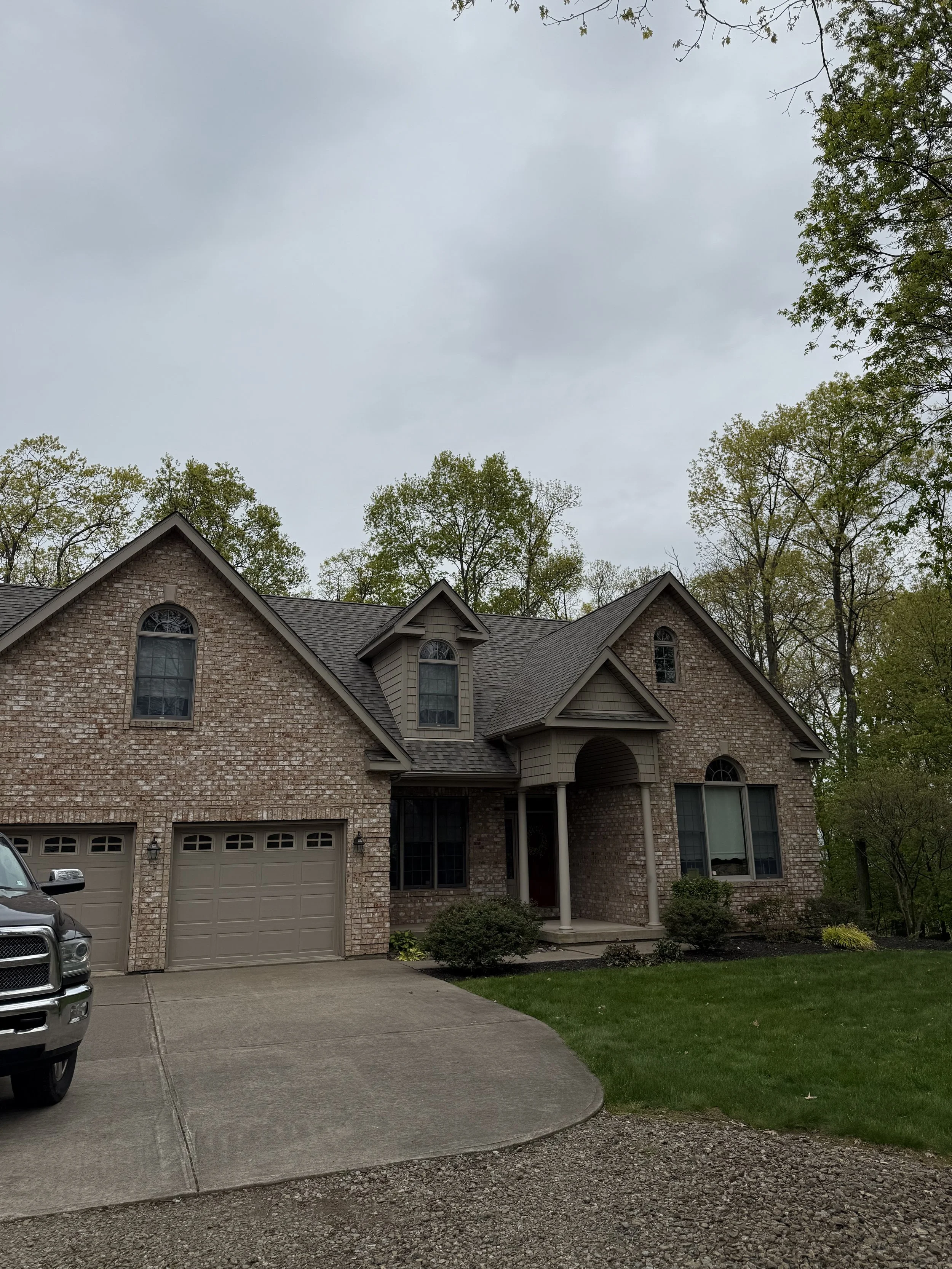 A two-story brick house with multiple gable roofs and arched windows. There is a driveway with a gray garage door, a black and silver truck parked on the left, and a lawn with small bushes and trees around the house. Overcast sky with gray clouds.