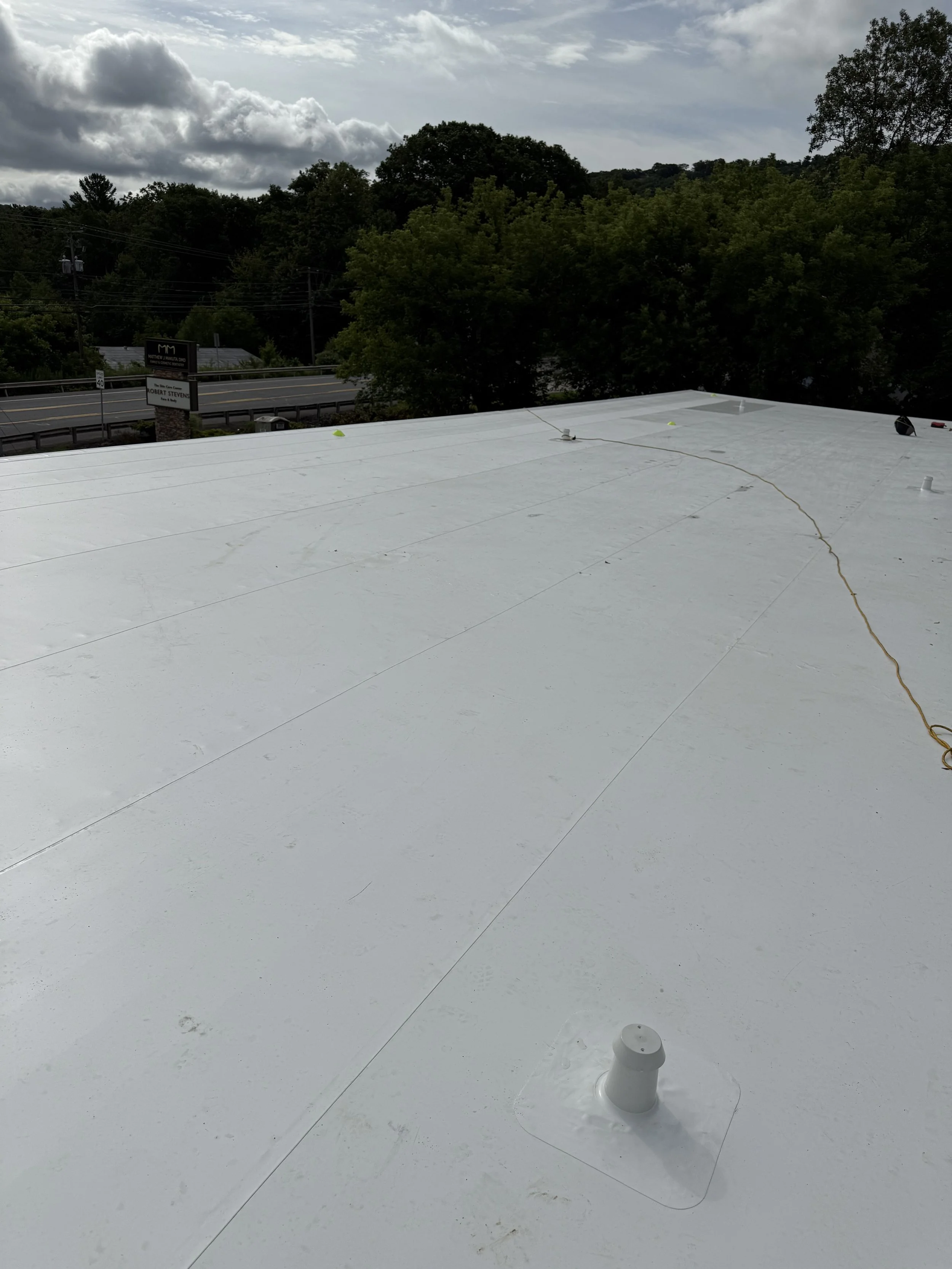 View of a flat white roof with some small vents and wires, overlooking a road, green trees, and a cloudy sky.