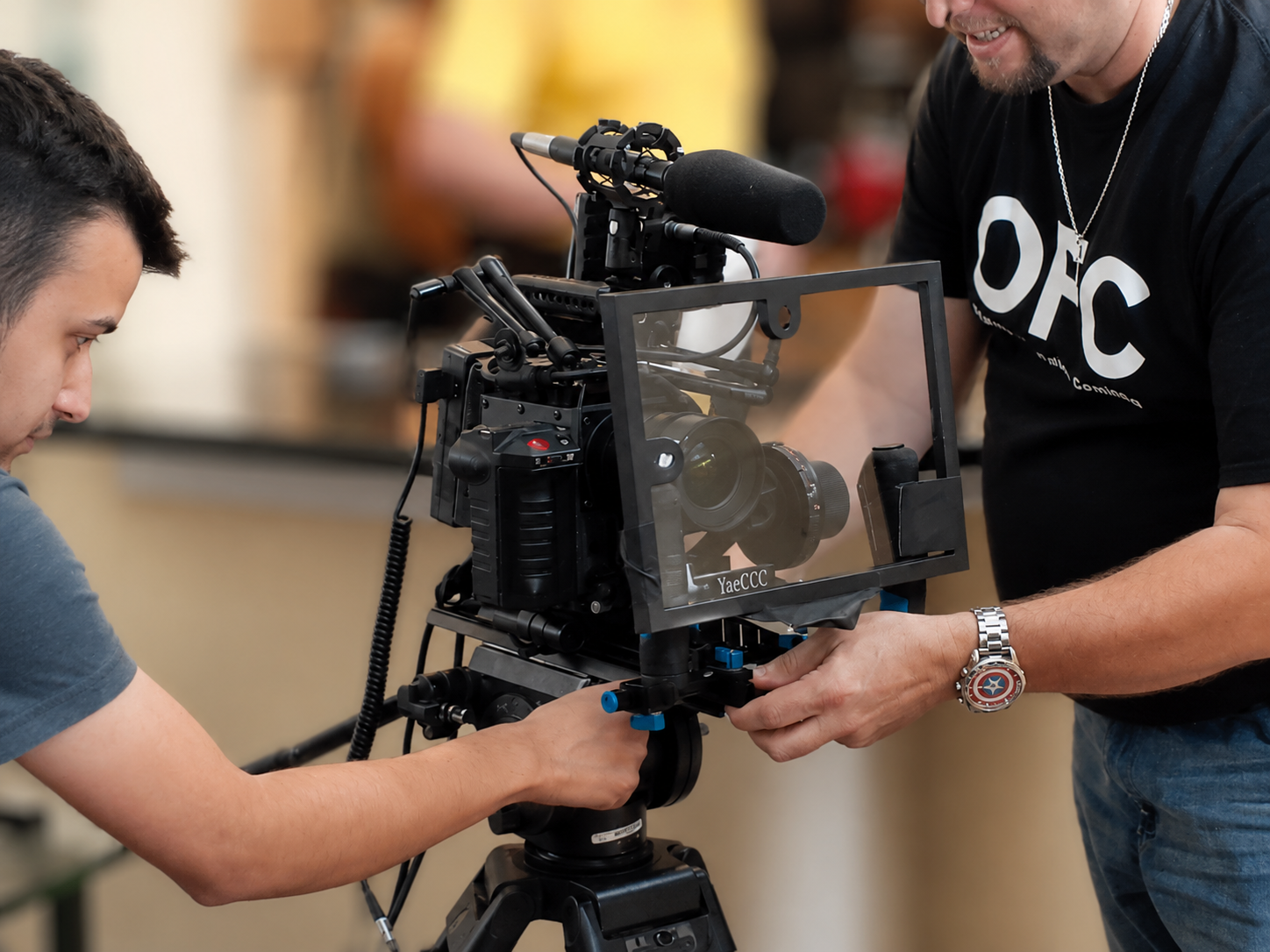 Two men are setting up a professional film camera, with one adjusting the camera on a tripod and the other assisting, in an indoor setting.