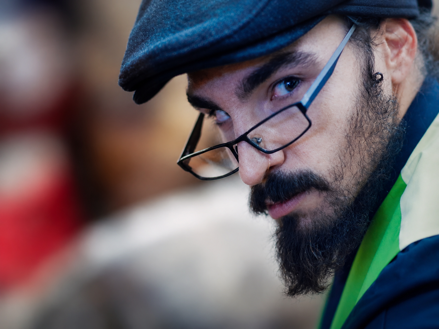 Close-up of a man with glasses, dark beard, and mustache wearing a cap and yellow safety vest, focused on something.