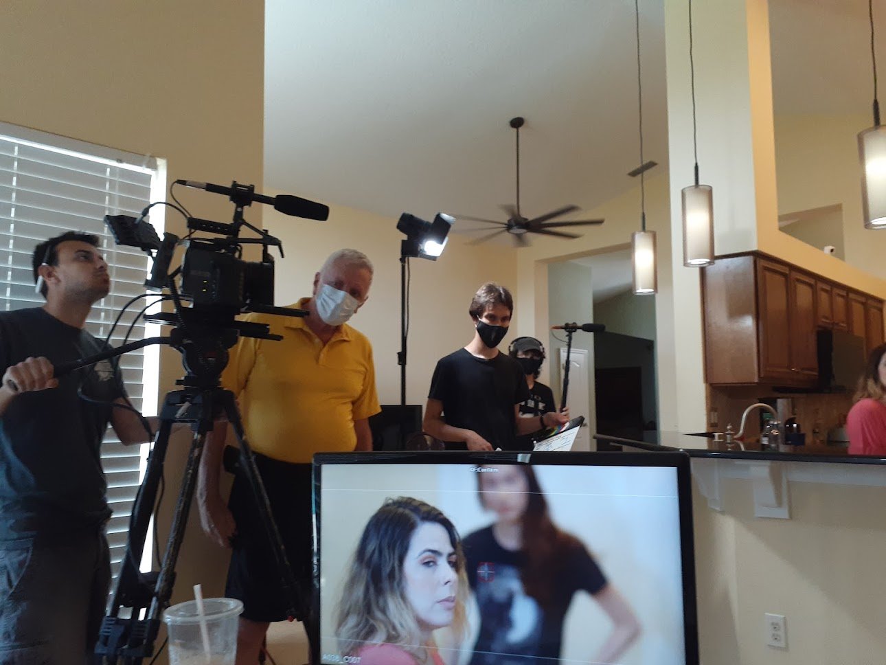 Filmmaking crew on set in a kitchen, with camera, lighting, and crew members, including a woman with long brown hair seen on a monitor in the foreground.