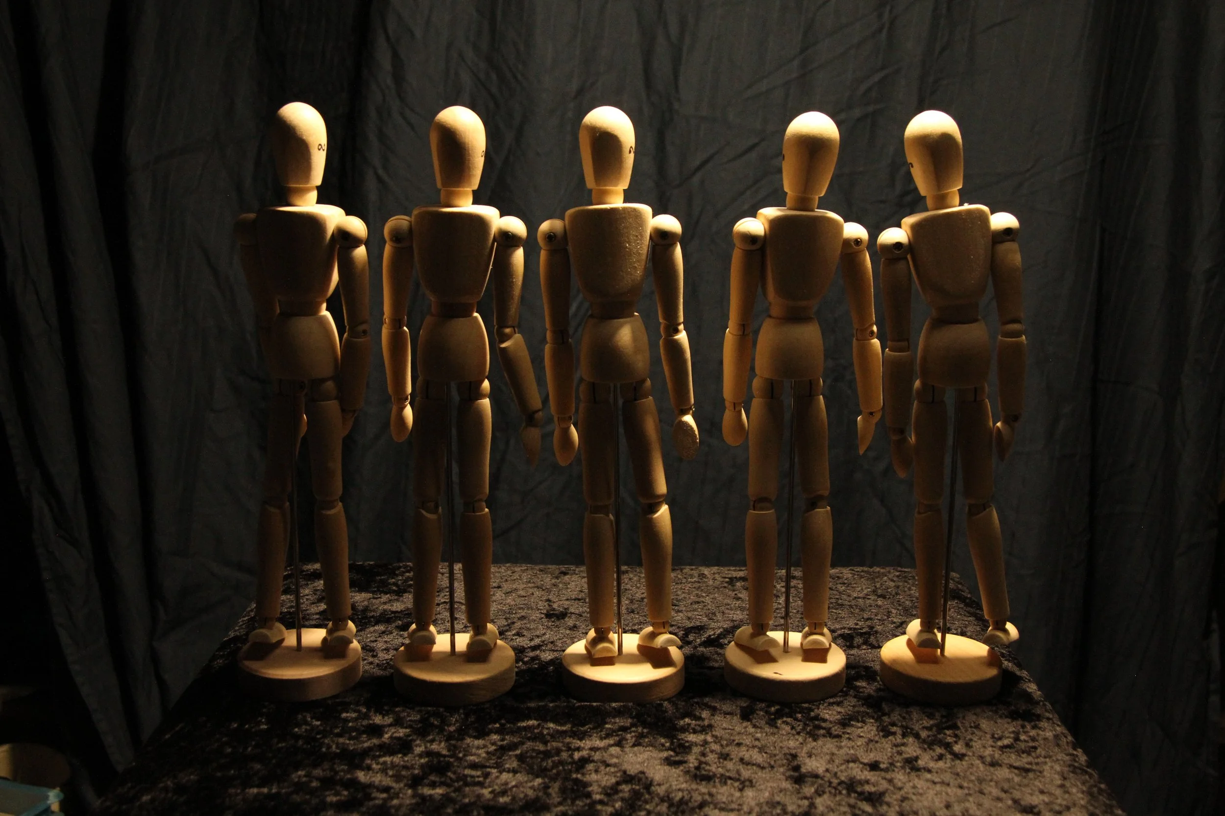Collection of wooden artist mannequins standing on black fabric with a dark background, illuminated by a single light source.