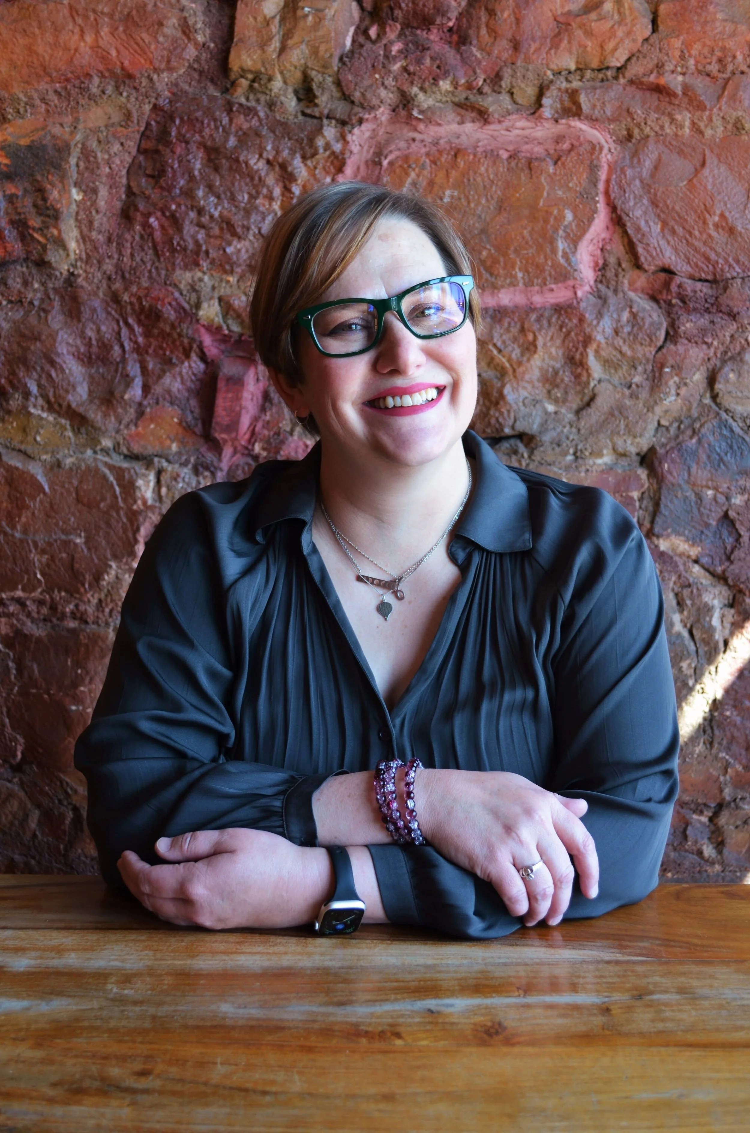 A woman with short brown hair and glasses smiling, sitting at a wooden table with her arms crossed in front of a red stone wall. She is wearing a dark gray shirt, jewelry, and a smartwatch.