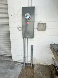 Electrical meter box mounted on a white wall, with two exposed electrical conduits running into the ground.