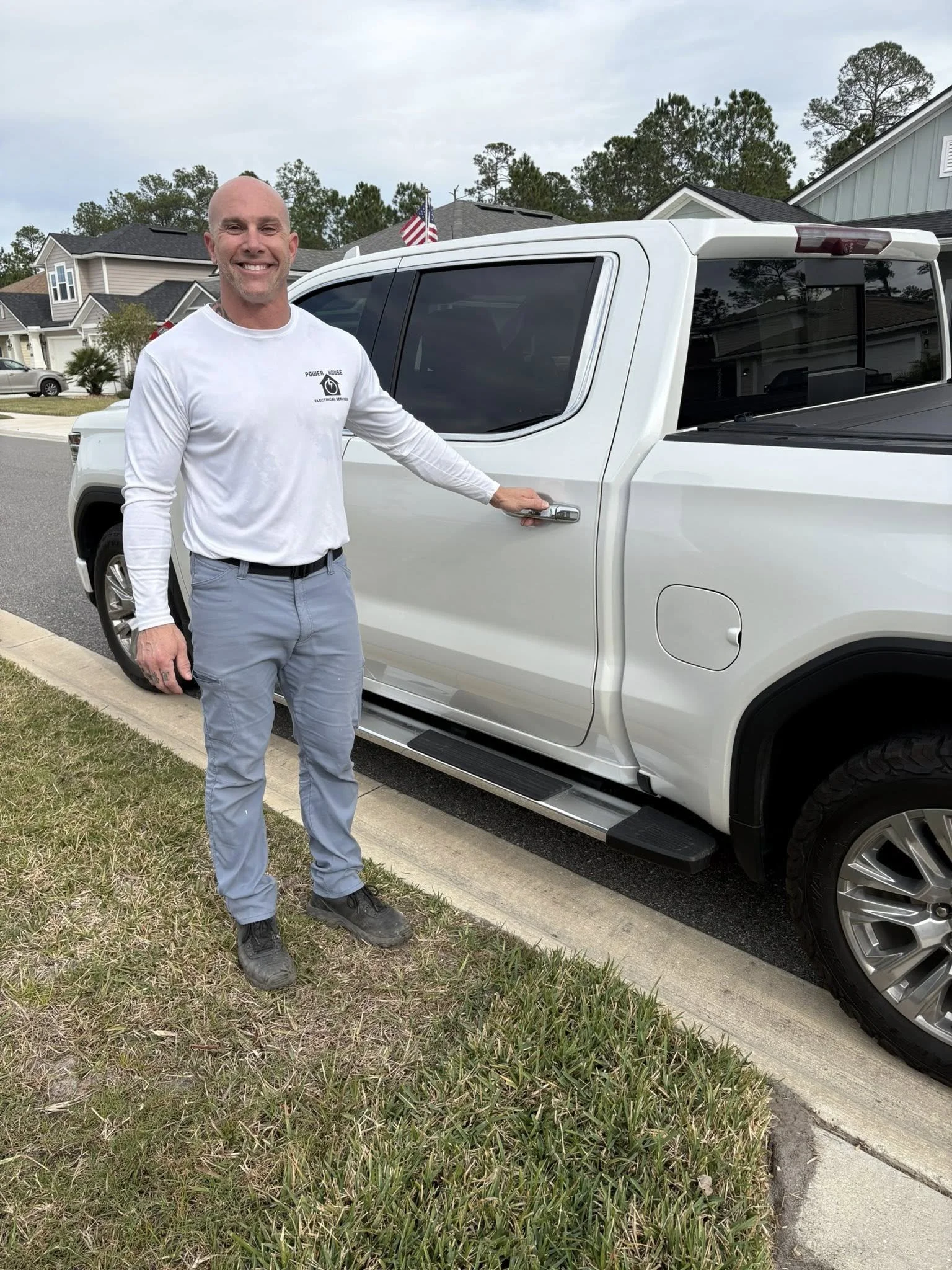 A smiling man in a white long sleeve shirt and light pants standing next to a white pickup truck parked on a residential street. The man has his right hand on the truck's door handle, and there are houses and trees in the background.