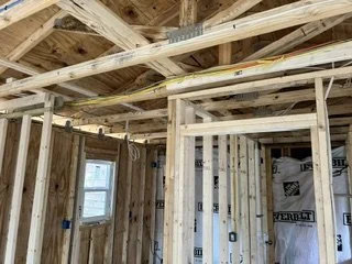 Interior view of a house under construction, showing exposed wooden framing, ceiling beams, and a window.