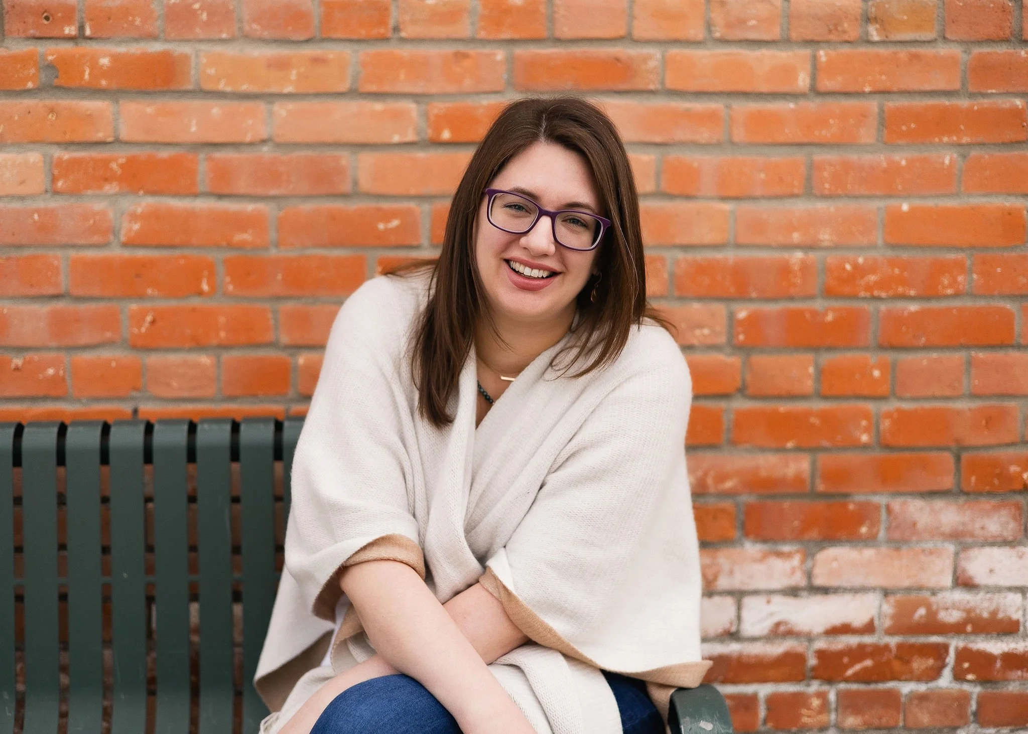 A woman with brown hair and glasses smiling, sitting on a bench against a brick wall.
